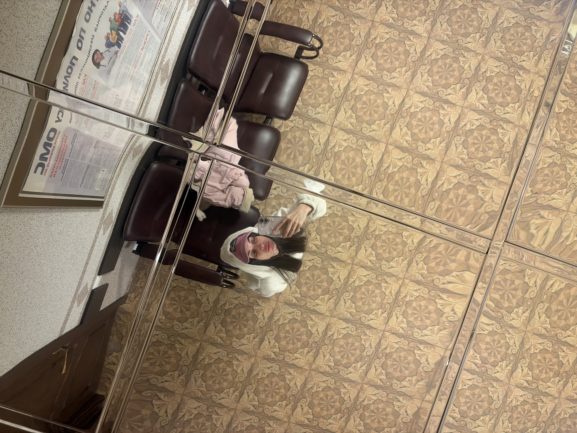 This image presents an unusual, inverted perspective of an indoor public space, captured as a reflection in a mirrored ceiling in Saint Petersburg, Russia. The ceiling comprises square, reflective tiles separated by bright metal strips, forming a grid pattern.
The central subject is a young woman, seen from above, looking upwards toward the camera. She has long dark hair and is wearing a white hooded sweatshirt or jacket, along with a pink baseball cap. She holds a smartphone up, partially covering her face, indicating she is taking a selfie or a reflective photo. Her expression is neutral.
The reflection also reveals several dark brown, padded chairs, characteristic of a waiting area in a public building. On the left wall, a large poster or notice board is visible in the reflection. Text on the poster includes Cyrillic words such as "КАК" (How), "ОАО", and possibly "СВОЙ КРУГ" (Own Circle). Latin characters, including "UR.ZMOBRZ.Www", also appear, suggesting a website or partial URL. The floor beneath the reflected elements features distinctive light brown patterned tiles.
The setting appears to be a public waiting room, possibly in a clinic, office, or transportation terminal, characterized by its functional seating and informational displays. The lighting is artificial, illuminating the scene. The image offers a unique, overhead view of the environment and its occupants through the mirrored surface.