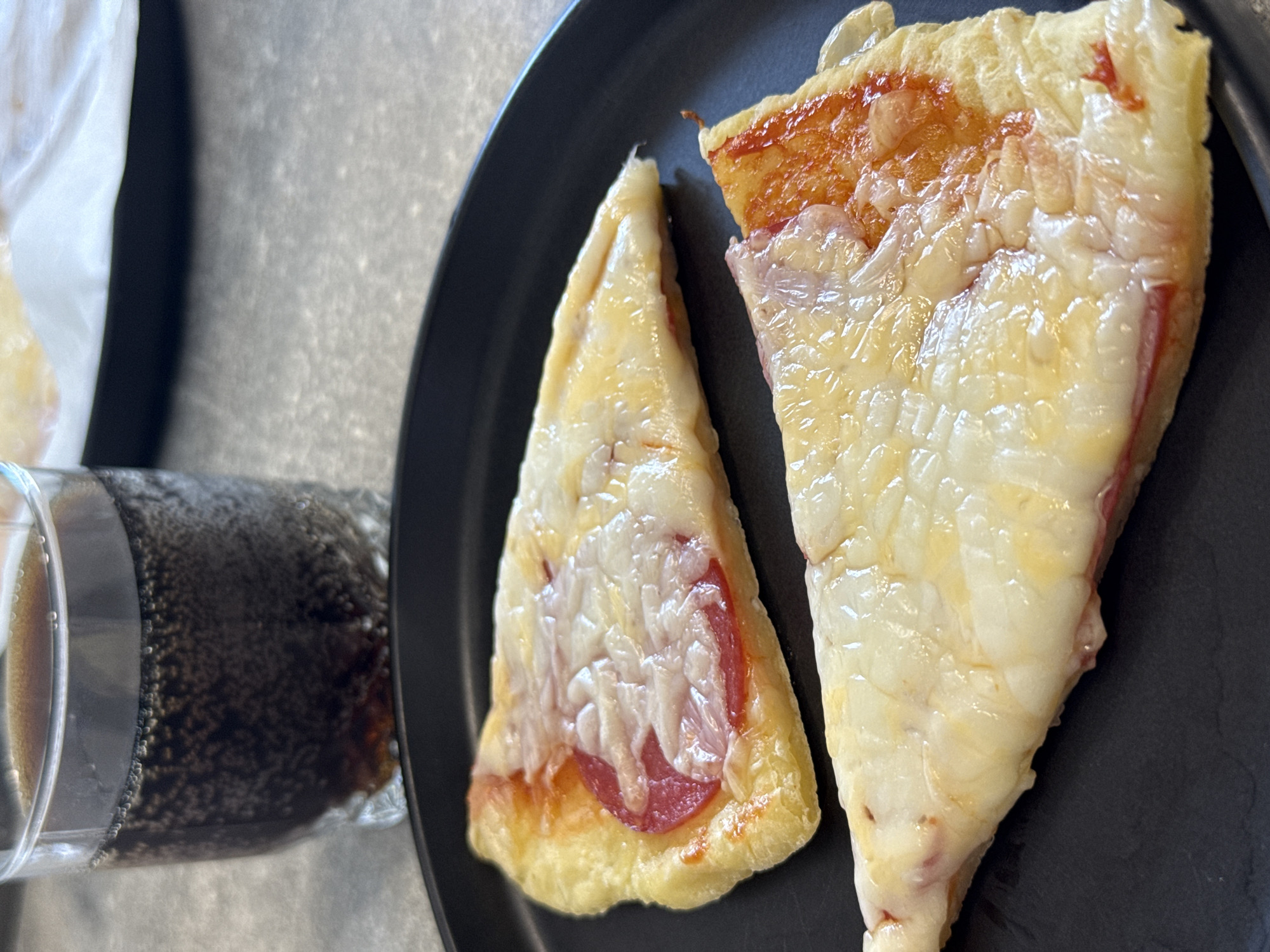 The image shows two slices of pizza on a black plate, with a glass of dark liquid filled with bubbles in the background. The pizza slices are triangular and covered in melted cheese. One slice has a visible piece of pepperoni. The glass contains a carbonated beverage, likely cola. The setting appears to be a kitchen or dining area with a grey countertop. There is no information in the image to suggest a location in Lyubertsy, Russia.