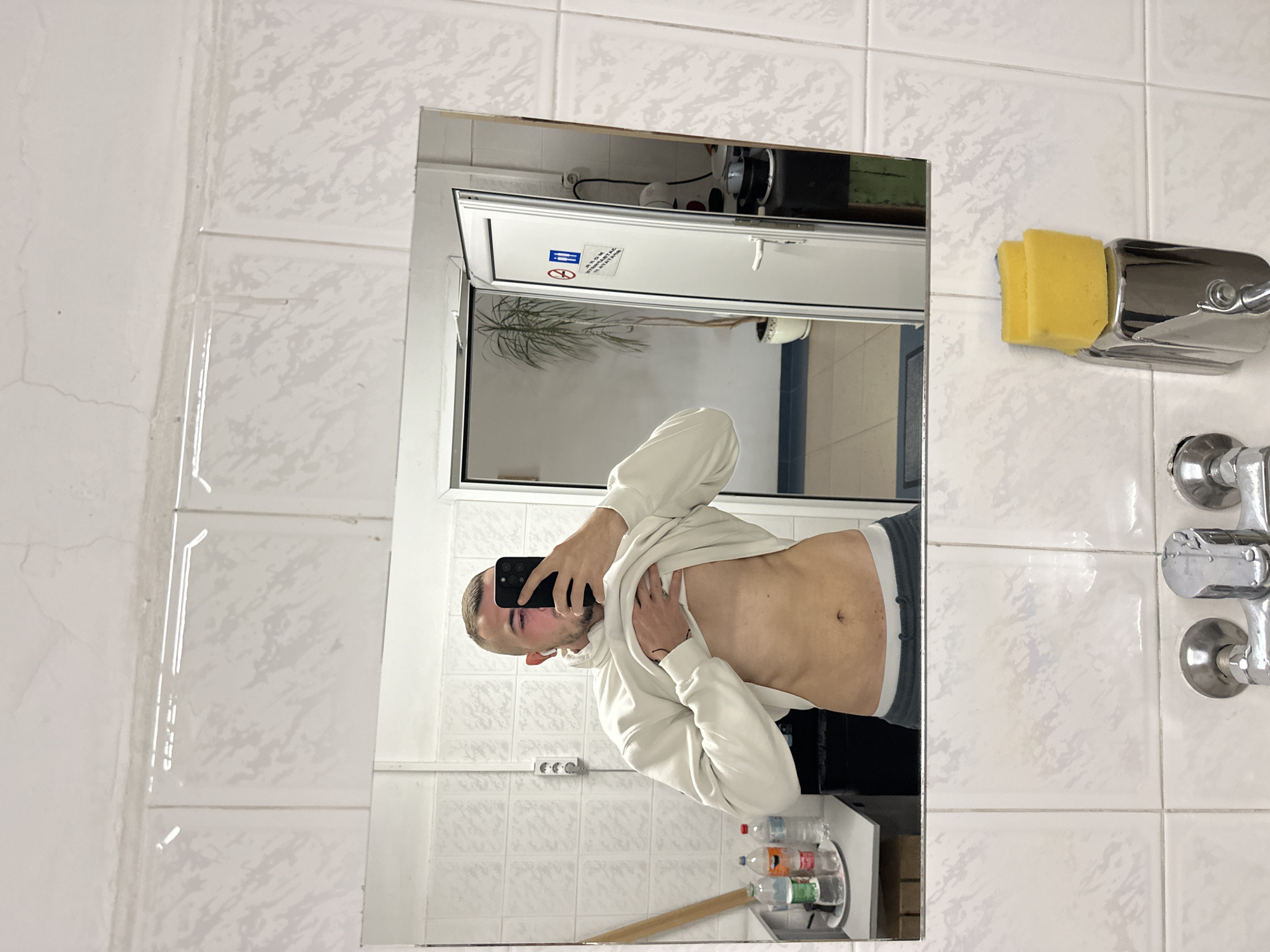 A male individual is captured in a mirror selfie, standing in what appears to be a tiled bathroom or changing room. He is wearing a light-colored hooded sweatshirt, which he has pulled up to expose his stomach and lower chest. He holds a black smartphone to take the picture, with an earbud visible in his left ear.
Within the mirror's reflection, behind the man, a white tiled wall with a three-socket electrical outlet is visible. Below, a metal rack holds various plastic bottles (water and soft drinks) and a wooden stick or handle. A white-framed door or window, with a glass panel, reflects a plant, possibly a palm. Above this frame, a gap reveals some wiring and a wooden beam.
Outside the mirror's frame, on the surrounding white tiled wall, there are signs of wear including stains and cracks. To the right, a chrome shower unit with two faucets is mounted, and above it, a chrome holder contains two yellow sponges.
On the door/window reflected in the mirror, a sticker displays Cyrillic text and symbols. Partially legible phrases include "ЗАБРАНЕНО ПУШЕНЕ" (No Smoking) and "МОЛЯ ПАЗЕТЕ" (Please keep), accompanied by a crossed-out cigarette symbol. The utilitarian aesthetic of the room, characterized by simple white tiles and exposed fixtures, suggests a functional or institutional environment. The image is presented rotated 90 degrees clockwise.