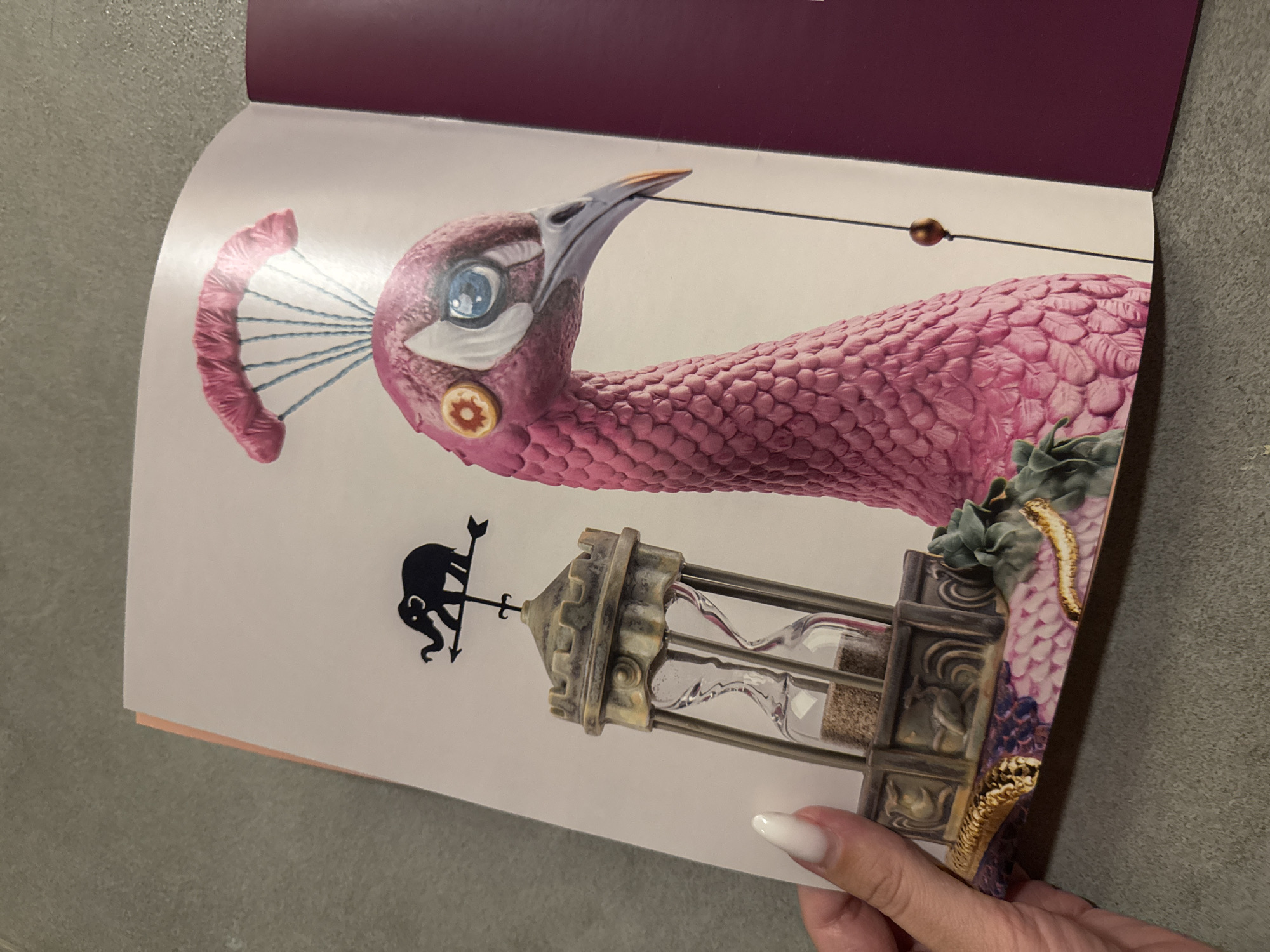 A close-up, high-angle shot captures a person holding an open book or magazine, revealing a surreal and intricate illustration on its right page. The background consists of a textured, grey surface, likely a floor or table.
The illustration depicts a fantastical pink, bird-like creature with a textured, scaly or feathered body. Its head features a sharp beak, a bright blue eye with a white outline, and a small, orange, gear-shaped embellishment on its cheek. A thin black line, threaded with a small golden bead, extends from the creature's beak towards the top right of the page. Attached to the back of its head is a pink, ridged, semi-circular object resembling a parachute or fan, connected by thin blue lines.
Below the creature, an ornate, stone-like tower structure houses two hourglasses; the upper one appears to contain a flowing liquid, while the lower one holds fine granules. Topping the tower is a weather vane featuring the silhouette of an elephant holding an arrow. Green foliage and a golden, snake-like form are intertwined around the base of the pink creature and the side of the tower.
In the bottom right corner, a hand with long, white-manicured fingernails holds the book open. The left page of the book is largely blank, while a dark maroon border is visible at the top of the open spread. No visible text is present.
