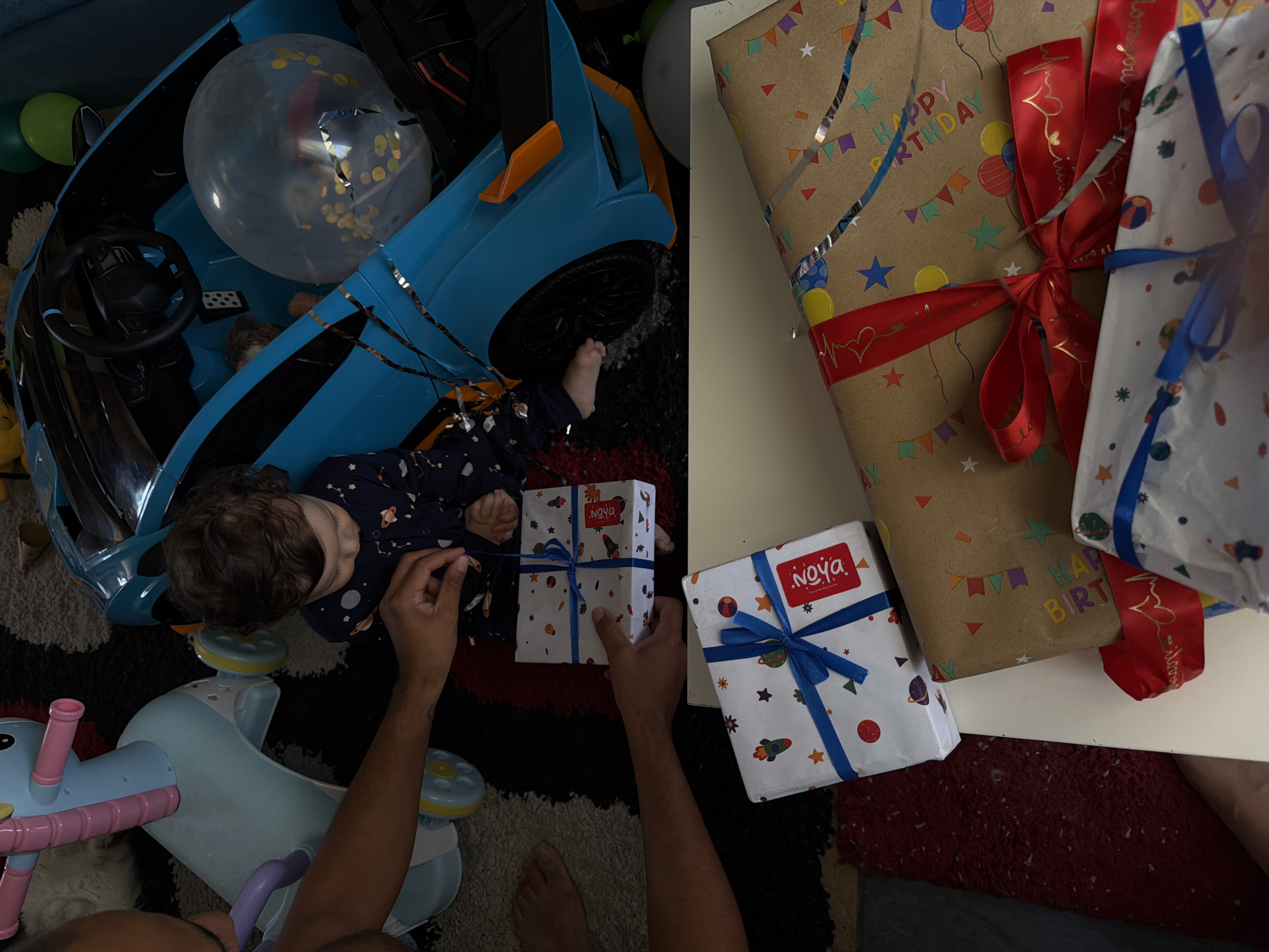 The image depicts a scene with a toddler in pajamas, sitting on the floor next to a blue toy car. A person's hands are visible, interacting with a wrapped gift in front of the toddler. Several wrapped gifts are present, some on the floor and others on a white surface. A unicorn-shaped ride-on toy is also in the background. The environment appears to be a domestic setting, possibly a living room or playroom, with a patterned rug. The presence of gifts and a toddler suggests a celebration, likely a birthday. There is no discernible visual information in the image to indicate a specific location in Sharm El Sheikh, Egypt.