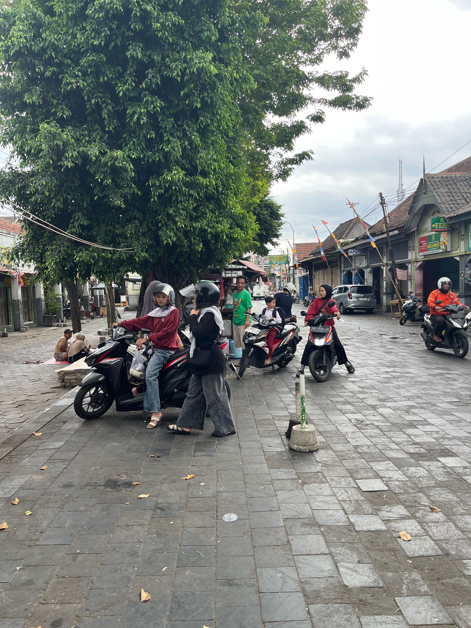 The image depicts a street scene in Yogyakarta, Indonesia, likely near a market or commercial area. Several people are present, most of whom are on or near motorcycles. In the foreground, two individuals, one wearing a maroon hoodie and jeans, and another in dark clothing and a headscarf, are standing by a black motorcycle. Both are wearing helmets. To the left, a person on a motorcycle is looking towards the camera, wearing a maroon hoodie, jeans, and a helmet. Further back, another person on a motorcycle, wearing a red hijab and black pants, is also looking ahead. In the background, there are buildings with traditional Indonesian architecture, some with signs advertising shops. Several other motorcycles are parked or being ridden by individuals, some wearing helmets and others not. The street is paved with dark gray tiles. A large tree with dense green foliage dominates the left side of the frame, casting shadows on the street. The sky is overcast with clouds. The overall atmosphere suggests a typical day in a bustling urban environment.