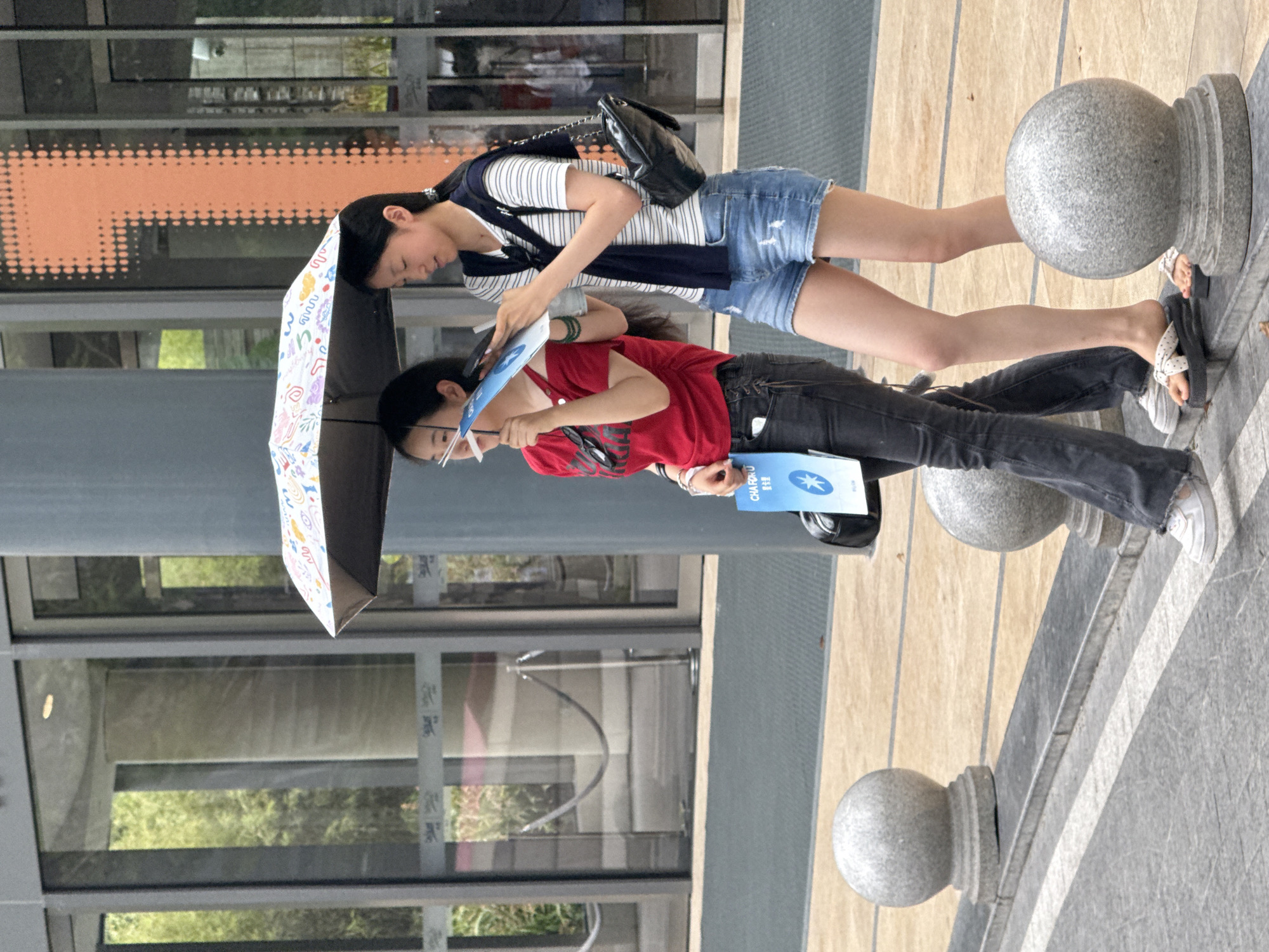 Two young women are walking outdoors under a colorful patterned umbrella. The woman on the left, wearing a red off-the-shoulder top and dark jeans, holds a blue pamphlet. The woman on the right, wearing a striped t-shirt, denim shorts, and a dark cardigan tied around her shoulders, is looking at a book or pamphlet. Both women are wearing casual footwear. They are walking on a paved area with decorative stone spheres along the edge. In the background, a modern building with large glass windows and an orange graphic design is visible. The scene appears to be in an urban environment, potentially a public space or promenade in Yuzhong District, China.