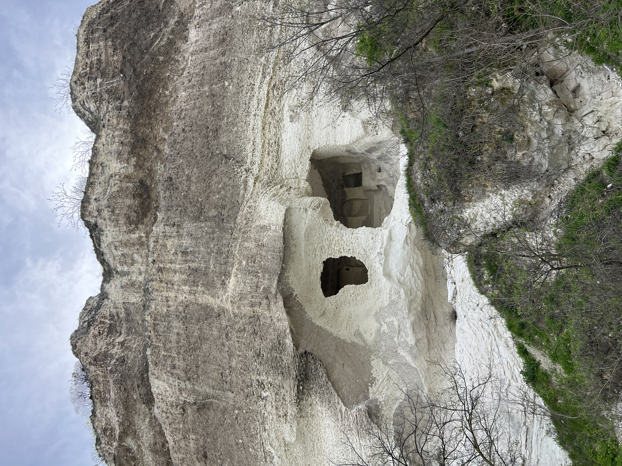 The image shows a rock formation in Nikopol, Bulgaria, featuring a rock face with a cave carved into it. The cave has multiple openings, giving the impression of rooms. The surrounding area has some sparse vegetation and bare trees. The sky is visible at the top left corner.