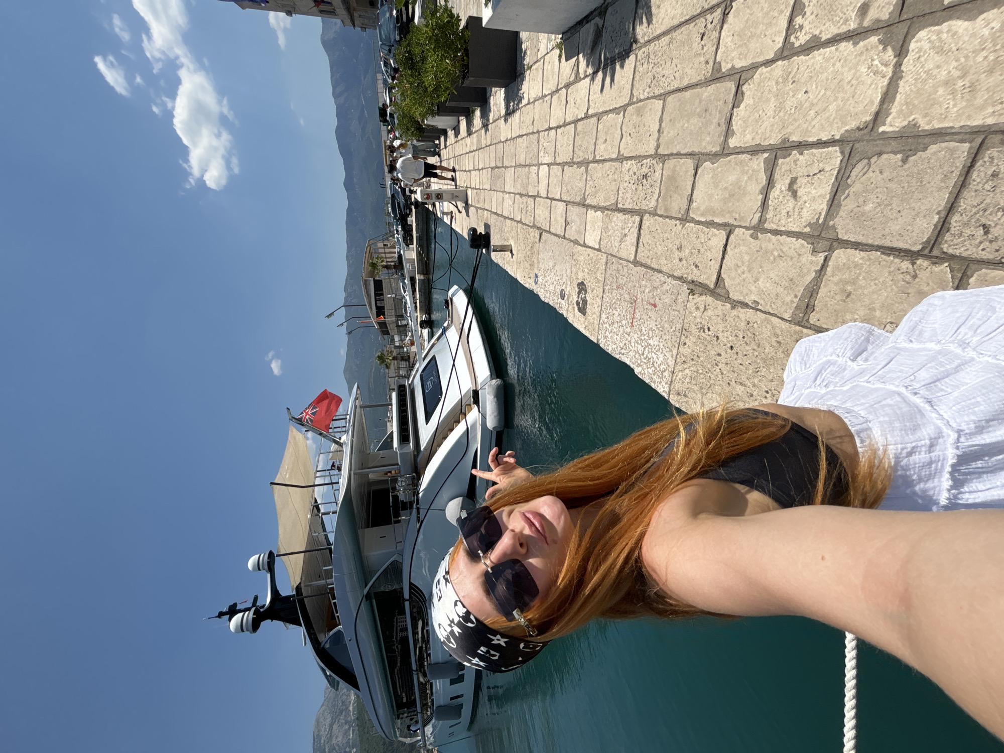 A woman with long red hair, wearing a black bikini top, a white skirt, a bandana, and sunglasses, poses for a selfie on a stone-paved pier in Budva, Montenegro. She is standing next to a large white yacht docked in the turquoise water. In the background, there are more boats, a street with parked cars, and mountains under a clear blue sky with scattered clouds. The yacht has a British flag flying from its stern. The scene depicts a sunny day in a harbor area.