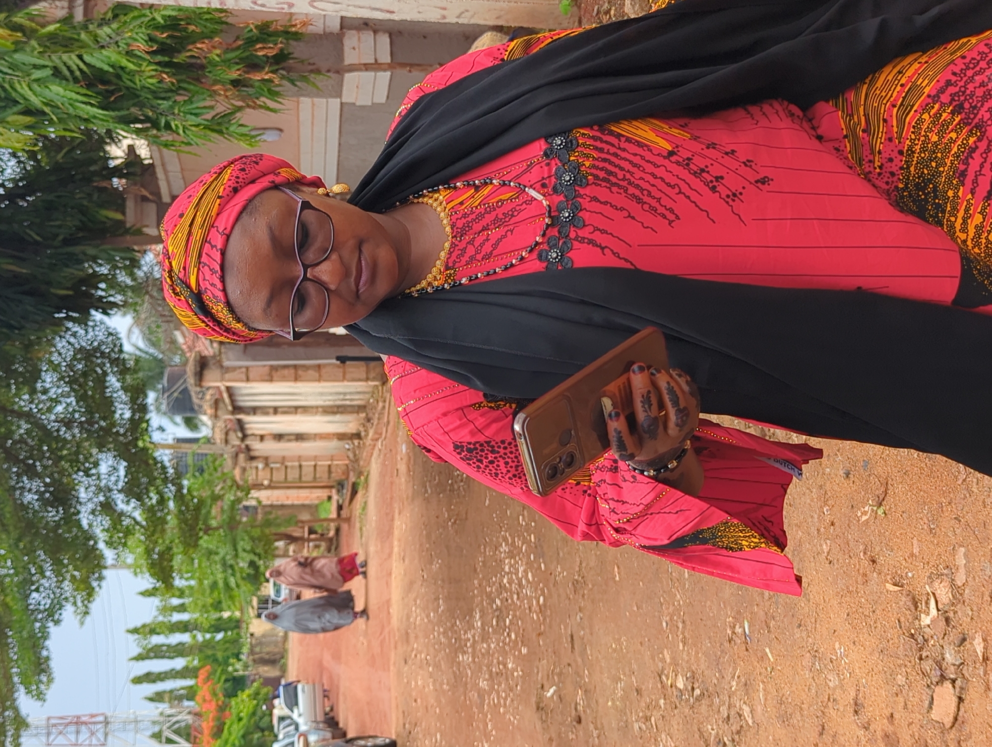 A woman is depicted standing outdoors on a sunny day in Unguwar Rimi, Nigeria. She is the primary subject, positioned slightly off-center and looking towards the viewer. She wears a vibrant, long-sleeved dress featuring a red background with intricate black and yellow patterns. A black headscarf or veil covers her head, and a black shawl is draped over her shoulders and upper body. She has glasses on, golden earrings, and two necklaces—one gold-toned and the other made of black and gold beads. Her hands are adorned with elaborate black henna designs, and she wears a simple bracelet on her right wrist. She holds a light brown smartphone in her left hand. Her expression is neutral.
The setting appears to be a semi-urban or rural area, characterized by a reddish-brown dirt path or road. Lush green trees and foliage are abundant, particularly to her left and in the background. In the middle distance along the path, two other individuals, also dressed in traditional long garments (one brown, one grey/blue), are walking away from the viewer. Behind them, buildings with light-colored pillars or columns are visible. Further in the background, parked vehicles can be seen, and a metal lattice tower structure is partially visible on the far left. The overall impression is daytime with clear weather. A small label on her dress, visible near her left hip, reads "DUTCH W".