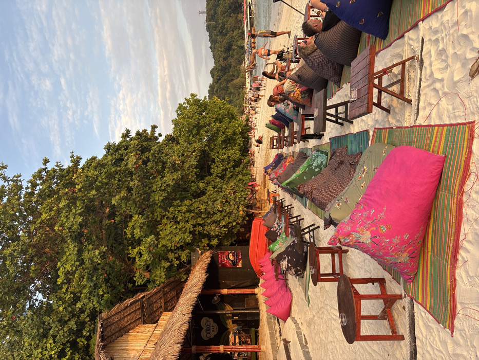 The image depicts a lively beach scene at Ban Ko Li Pe, Thailand, during golden hour. On the left, a wooden structure with a thatched roof, identified as "Monkey Bar," features an entrance flanked by bright pink bean bag chairs and a red inverted boat. Large green trees with thick foliage line the background behind the bar.
The foreground and mid-ground consist of rows of open-air seating arrangements on white sand. Each setup includes a colorful, patterned bean bag cushion placed on a striped mat, accompanied by a small, dark wooden table, some featuring ashtrays. Several individuals are seated on these bean bags, relaxing and engaging in casual interactions.
Towards the right and further down the beach, a larger group of people is seen walking along the water's edge. Some are in swimwear, others in casual attire, strolling along the shoreline. The calm, blue-green ocean in the background features several traditional longtail boats moored offshore. Beyond the beach and water, a lush, green hill rises, topped by a slender tower-like structure. The sky above is light blue with scattered cirrus clouds. The scene conveys a relaxed, evening atmosphere with warm, diffused lighting.