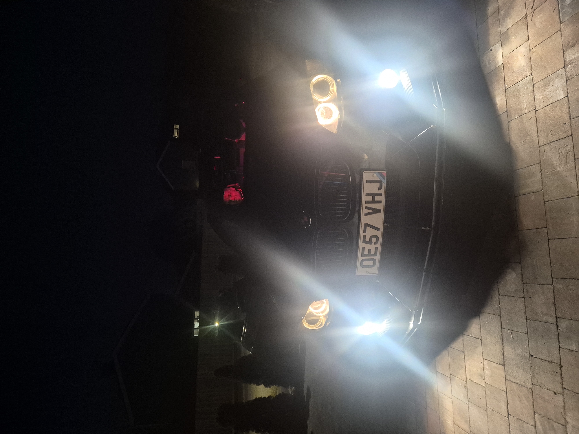 The image depicts the front of a dark-colored car with its headlights on, illuminating a paved surface. The license plate reads "OE57 VHJ". In the background, there's a house and some vegetation. The scene appears to be at night, likely in a residential area within West Suffolk, United Kingdom.