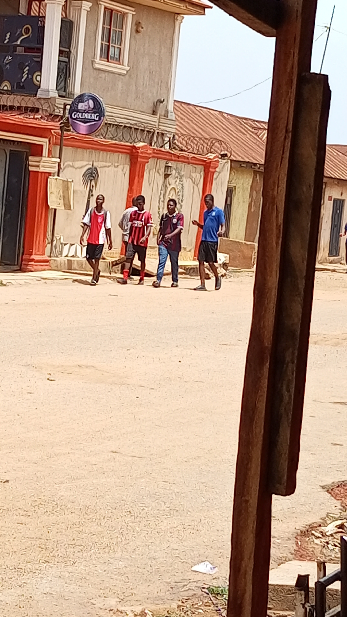 In Sabon Tasha, Nigeria, a group of four males is walking on a dirt road. All appear to be young adults. The first male is wearing a red and white soccer jersey with black shorts. The second male is wearing a red and black striped soccer jersey with black shorts. The third male is wearing a dark red and purple soccer jersey with blue jeans. The fourth male is wearing a blue t-shirt with black shorts. Behind the group is a building with a "GOLDBERG" sign. A wooden structure is in the foreground. The sky is overcast.