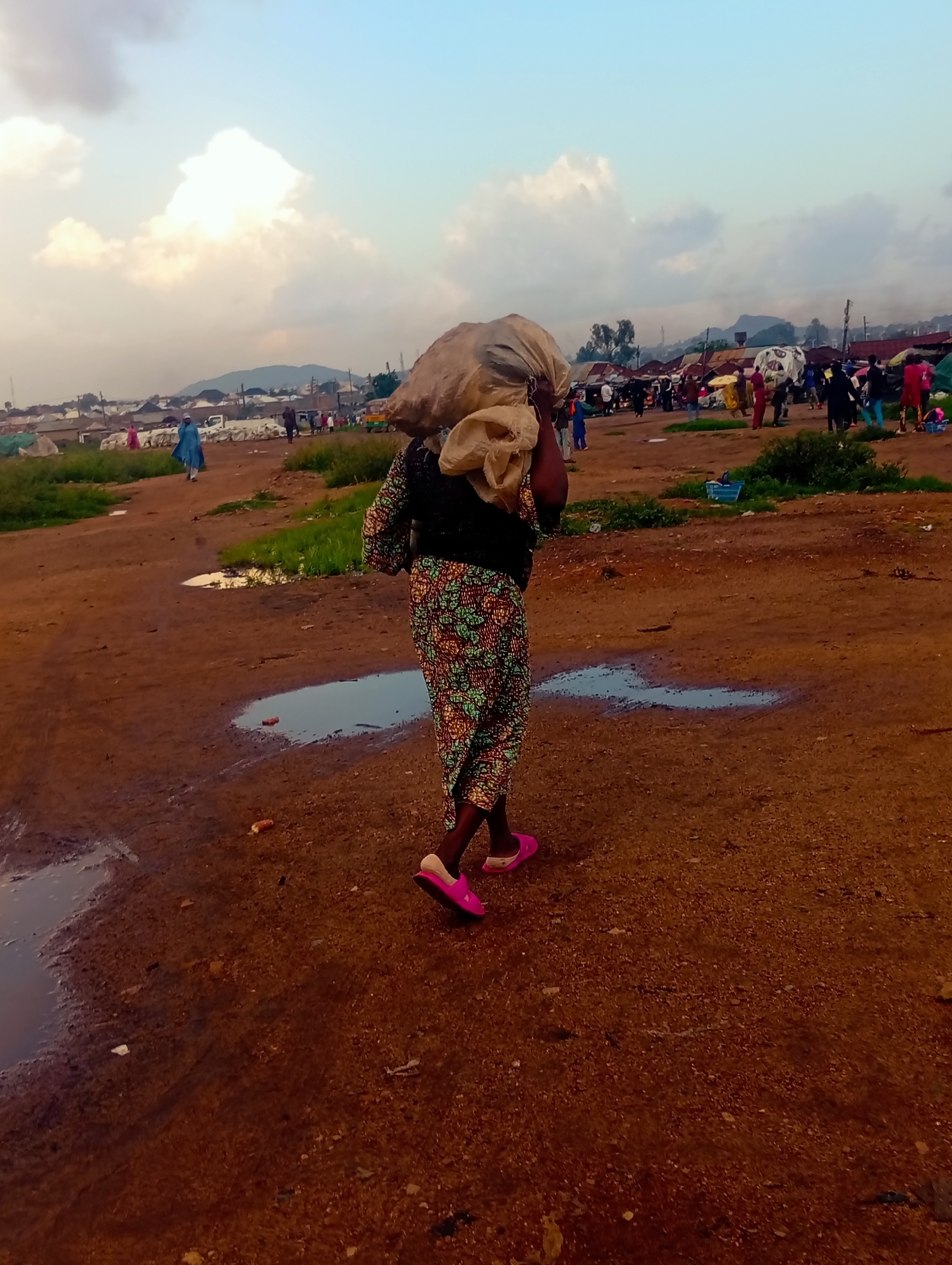 In this media file, a person walks away from the camera, carrying a large, light-colored bundle on their shoulders. They are wearing a patterned dress and bright pink slip-on shoes. The ground is a reddish-brown dirt with several puddles of water. In the background, a bustling marketplace is visible, with numerous stalls and a crowd of people. Some of the structures appear to be makeshift tents or shelters. In the distance, hills can be seen under a sky with soft, scattered clouds. The lighting suggests it is either early morning or late afternoon. The scene depicts everyday life in a busy outdoor market, possibly in Bukuru, Nigeria, based on the provided location context. There is no visible text in the image.