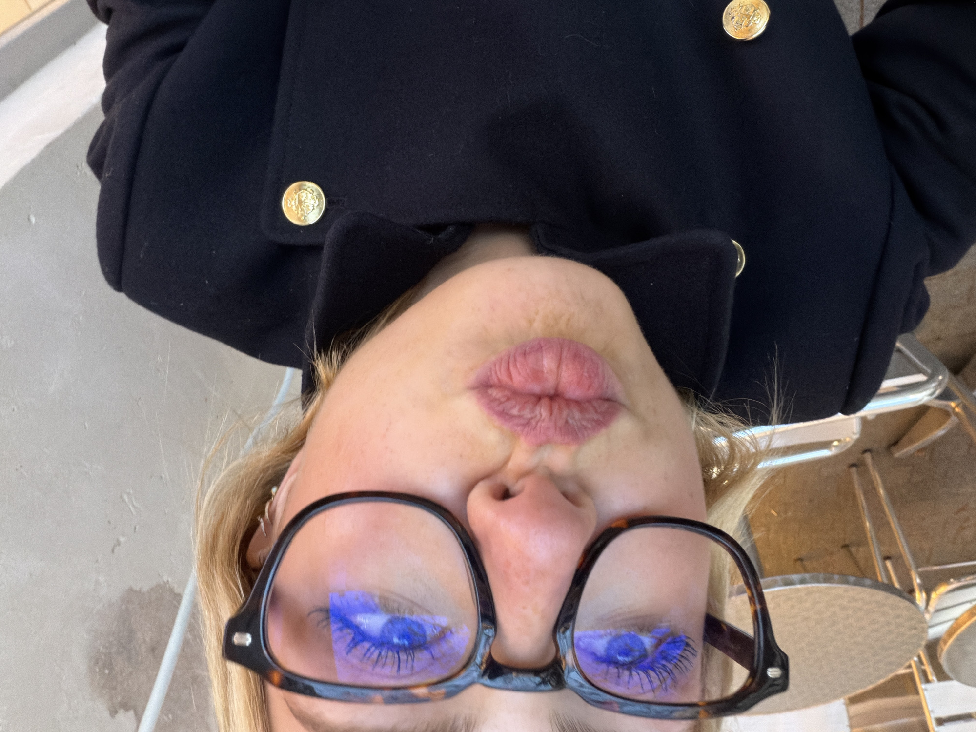 A close-up selfie features a light-skinned individual, likely female, with blonde hair framing the face. Freckles are visible on the nose. The individual wears dark tortoise-shell framed eyeglasses, with a distinct blue light reflection visible across the lenses, making the eyelashes appear blue. The lips are pursed into a prominent pout. The subject is wearing a dark, possibly navy or black, double-breasted peacoat or similar jacket, adorned with large, ornate gold buttons, each embossed with a crest or emblem. A small hoop or stud earring is visible on the subject's right ear. The background is out of focus, indicating a shallow depth of field. To the left, a portion of a round, metallic café table and a chair's back are discernible. To the right, a textured light-colored wall, possibly stucco or concrete, is visible, with a white vertical element (likely a pipe or railing) running along it. The scene depicts an outdoor café environment in Paris, France.