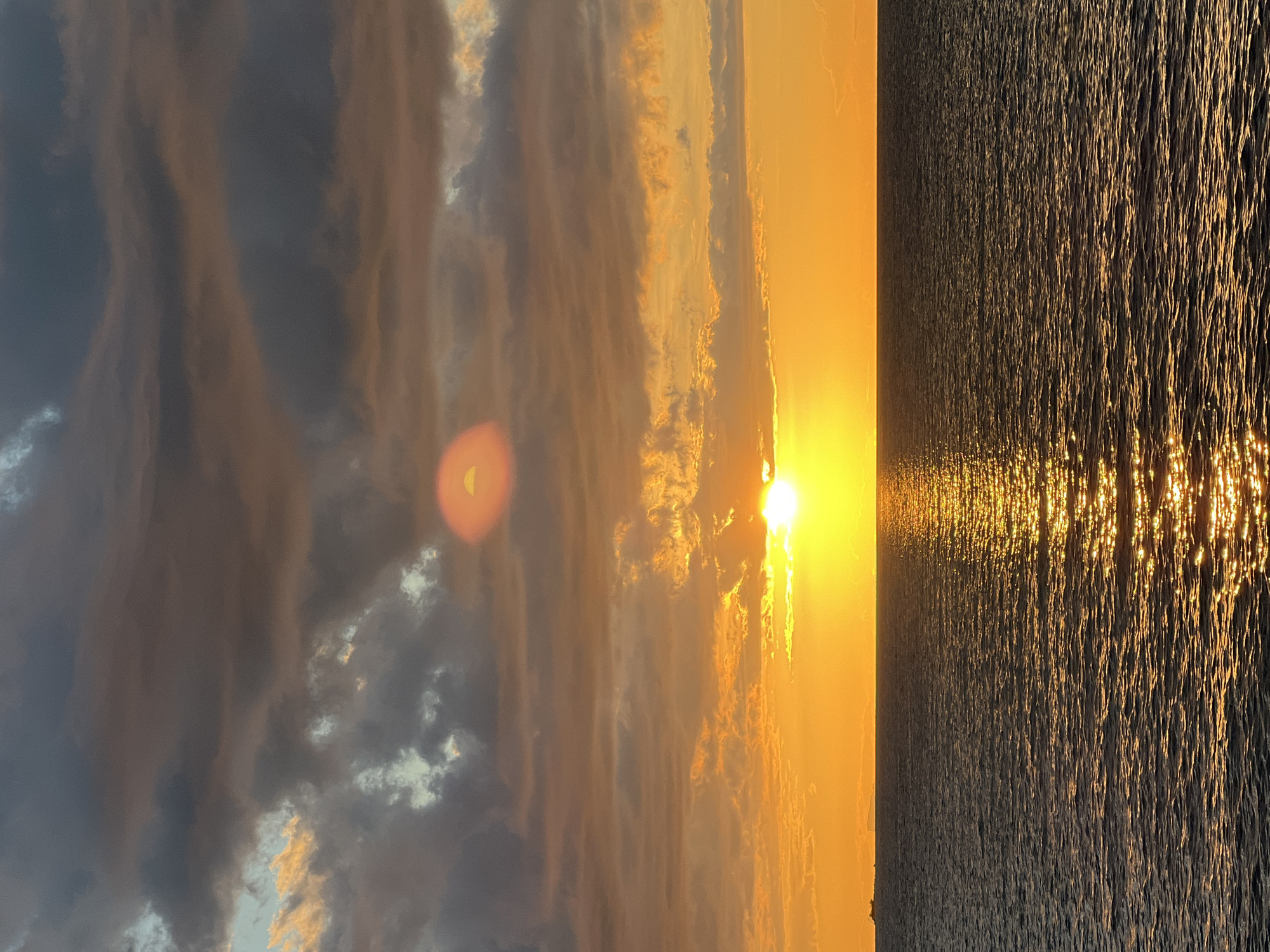 The image depicts a seascape at sunset, with the sun positioned low on the horizon, casting a warm orange glow across the sky and reflecting brilliantly on the rippling ocean surface. Dark, dramatic clouds with hints of orange and grey dominate the upper portion of the frame, partially obscuring the sun in one area and creating a lens flare effect. The sea occupies the lower half of the image, its dark, undulating surface shimmering with golden light from the sunset. A distant ship and a faint landmass are visible on the horizon, to the left of the setting sun. The overall scene is one of natural beauty and tranquility. This image was taken at Baan Plai Laem, Thailand.