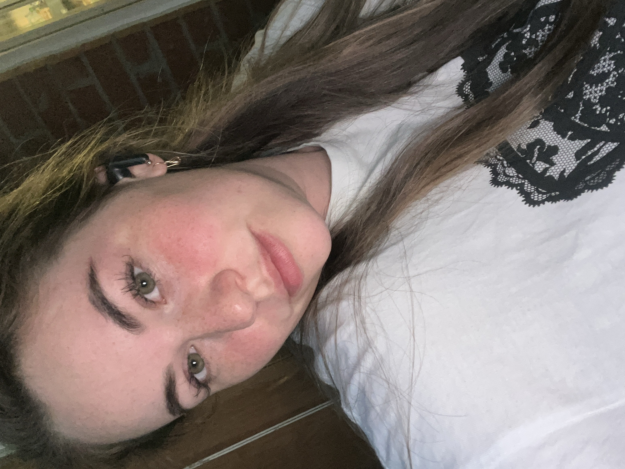 A young woman with long brown hair, green eyes, and fair skin is lying on her side, facing the camera. She is wearing a white shirt with black lace detailing on the shoulder. The background includes a brick wall and what appears to be a wooden surface. The image does not provide sufficient information to determine if the location is in the city of Chaikovsky, Russia.