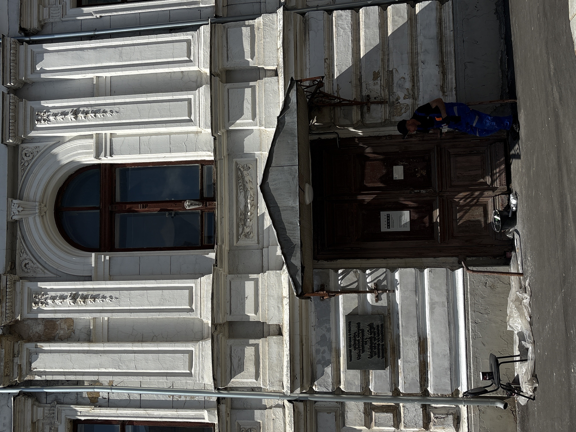 The image depicts the facade of a building with classical architectural elements, suggesting it is an older structure. The building is painted white and shows signs of wear and tear, with peeling paint visible in several areas. Ornate moldings and decorative elements adorn the exterior, including columns and a large arched window. Below this window, a dark wooden door is centered, with a small awning over it. To the left of the door, a black metal chair is placed on the sidewalk, next to a white plastic sheet and a black cup. A plaque with Cyrillic text is mounted on the wall to the left of the entrance. To the right of the door, a man dressed in blue overalls and a dark cap is leaning against a wooden pole, holding a phone. He appears to be taking a break. The sidewalk in front of the building is paved and appears somewhat worn. The overall scene suggests a public space in Moscow, Russia, possibly near a theater or historical building, given the signage on the door.