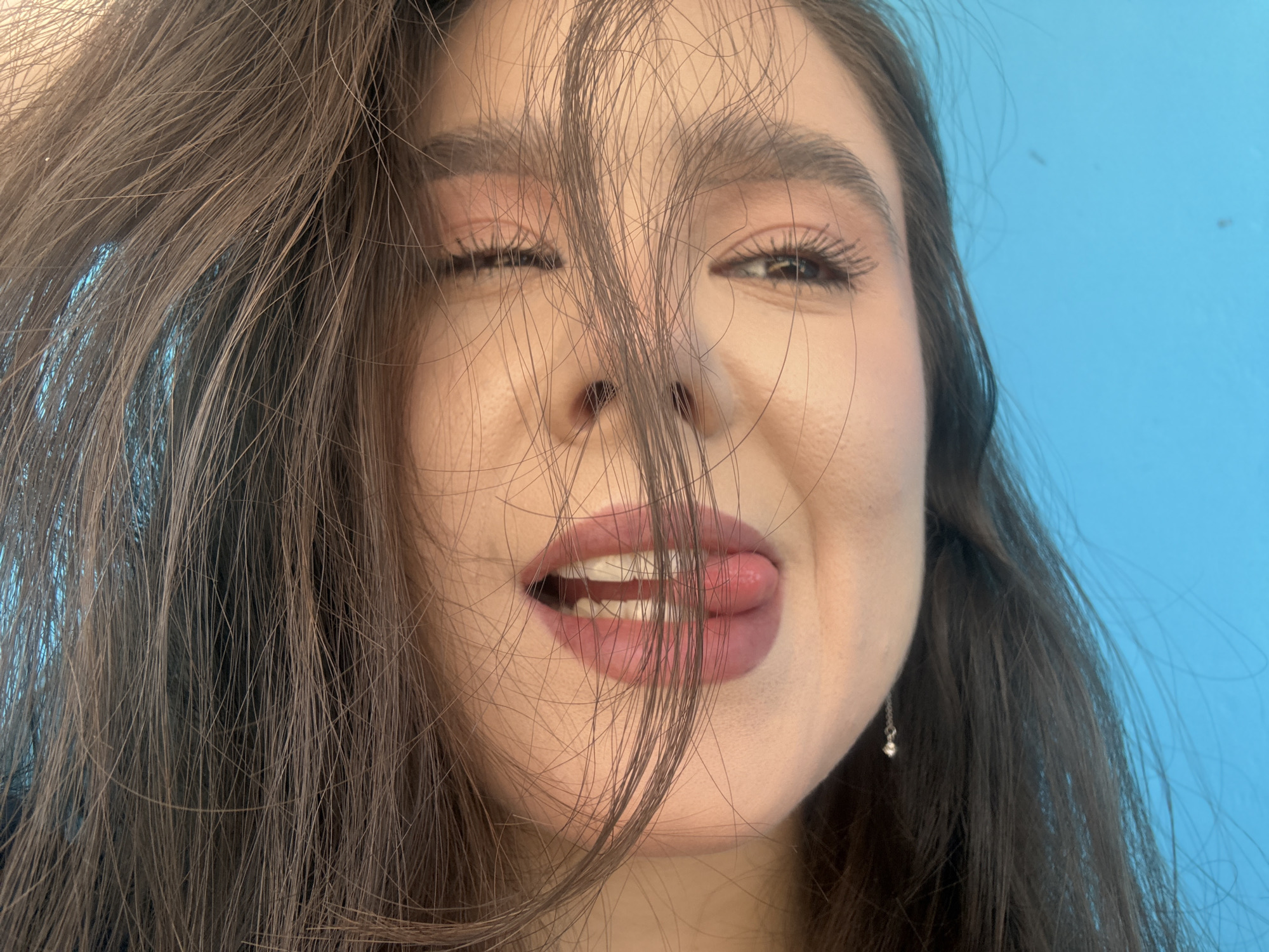 A close-up shot shows the face of a young woman against a bright blue background. Her dark hair is styled casually, with some strands falling across her face. She is looking towards the camera with a playful expression, her tongue sticking out slightly from her lips. Her eyes are partially closed, and she is wearing subtle makeup. A small silver earring dangles from her right ear. The lighting is bright, highlighting her features. The location is specified as Shulhynka, Ukraine.