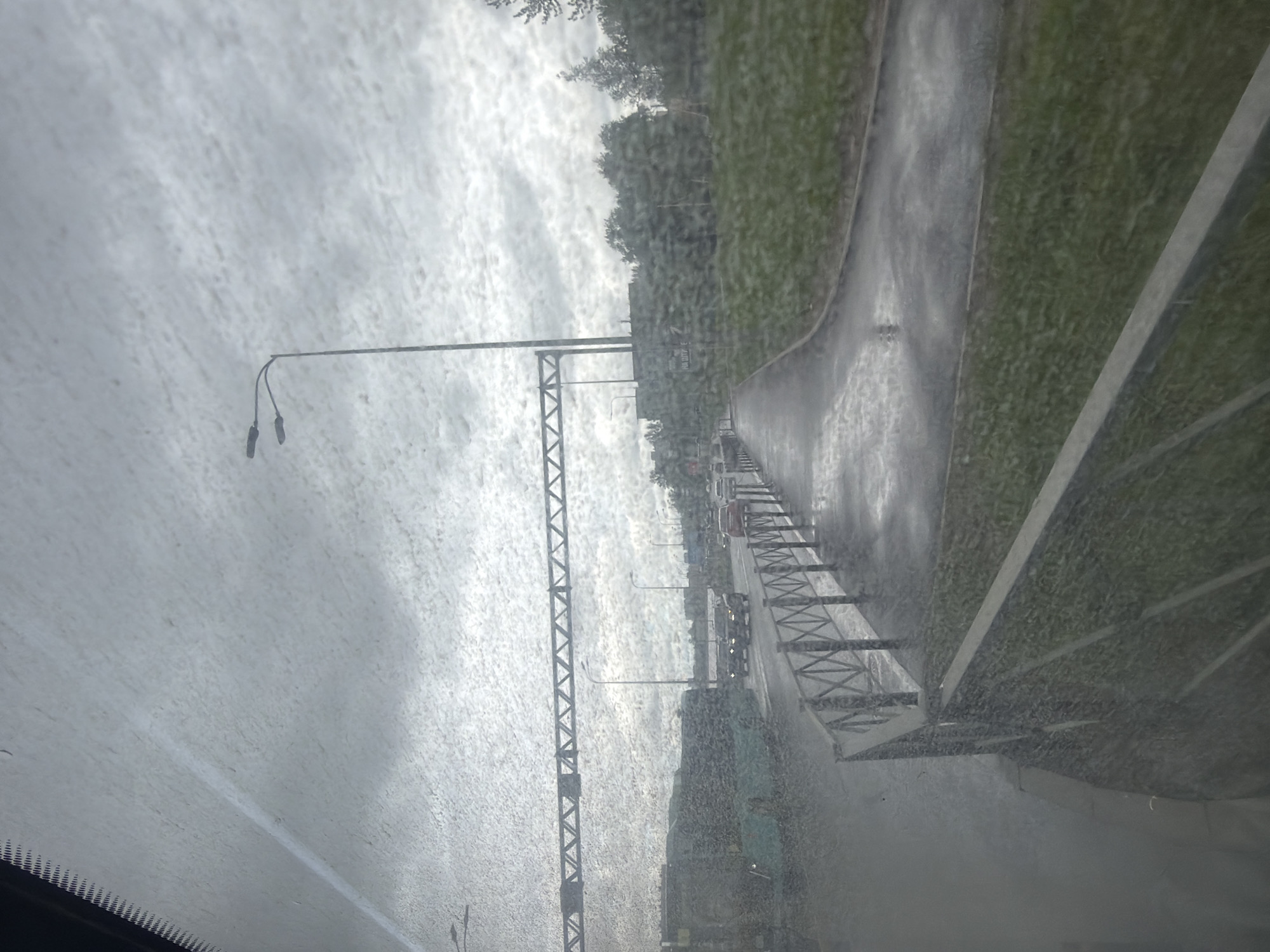 The image depicts a view from inside a vehicle, likely a car, on a rainy day. Raindrops are visible on the windshield. The scene appears to be a road, possibly a highway or major street, with a green area on one side. A metal structure, possibly part of a bridge or overpass, is prominent in the foreground. Streetlights are visible along the road. In the distance, there are cars and trucks driving on the road. Based on the architectural style of the bridge and other elements, the image is likely taken in the city of Saint Petersburg, Russia. The rain obscures details, making it challenging to identify specific locations or objects precisely.