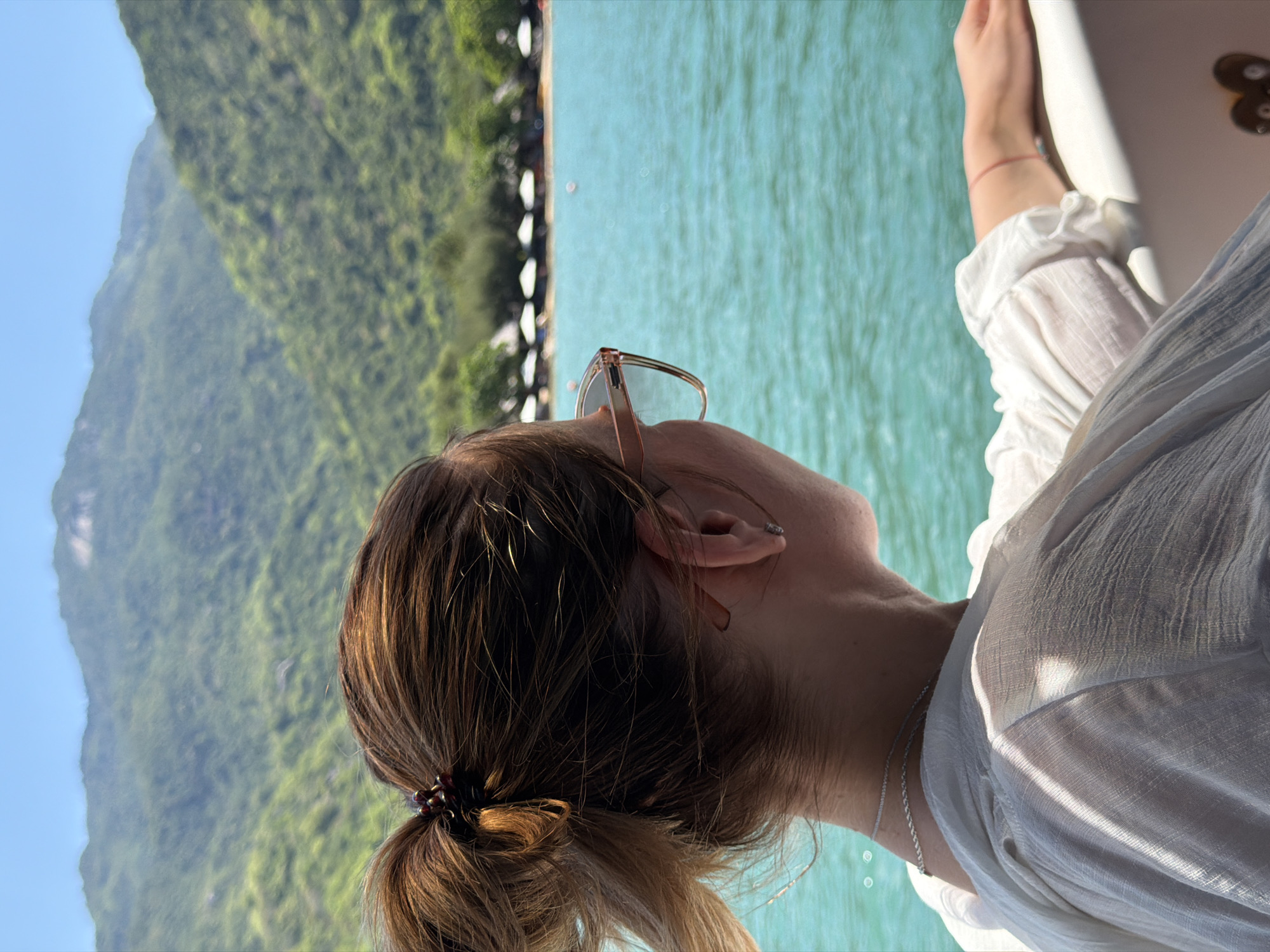 A woman with brown hair pulled into a ponytail, wearing a white top and sunglasses, is looking out at the ocean in Hoa Thang, Vietnam. The ocean is a vibrant turquoise color, and in the distance, green mountains rise against a clear blue sky. A beach with umbrellas is visible along the shoreline. The woman is leaning on a railing, her arm extended.