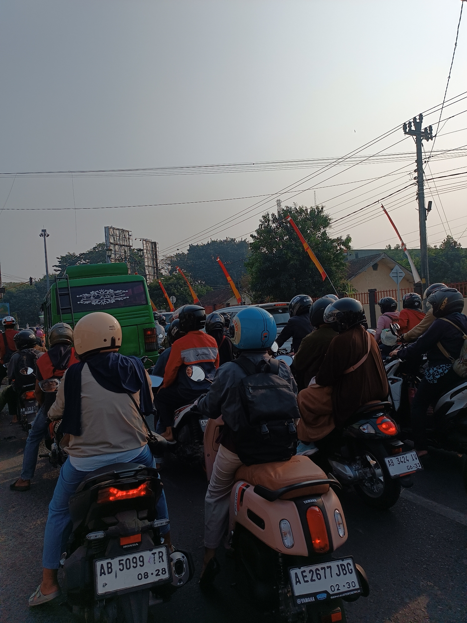 The image depicts a traffic jam of motorcycles in Banguntapan, Indonesia, on a street with overhead electrical wires and utility poles. In the foreground, several motorcyclists are visible, wearing helmets and casual clothing. One motorcyclist in the center wears a light-colored helmet and a blue scarf. Behind them, another rider wears a blue helmet and a black backpack. Further back, a green bus with ornate white lettering on its rear window is a prominent feature. The street appears to be asphalt, and there are buildings and trees in the background, suggesting an urban or semi-urban environment. Red and yellow flags are seen along the roadside, possibly indicating a local event or cultural display. The overall impression is one of a busy, sunlit afternoon in a Southeast Asian city.