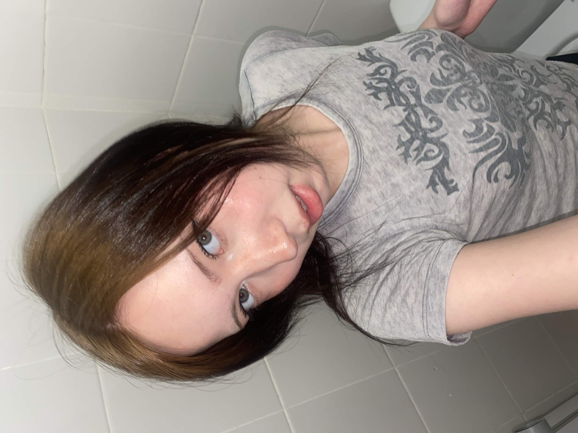 A young person with shoulder-length brown hair featuring blonde highlights is shown in a close-up, tilted headshot. The person is wearing a gray t-shirt with a dark gray ornate design. The background consists of white tiled walls and a portion of a white toilet. The location is presumed to be a bathroom.