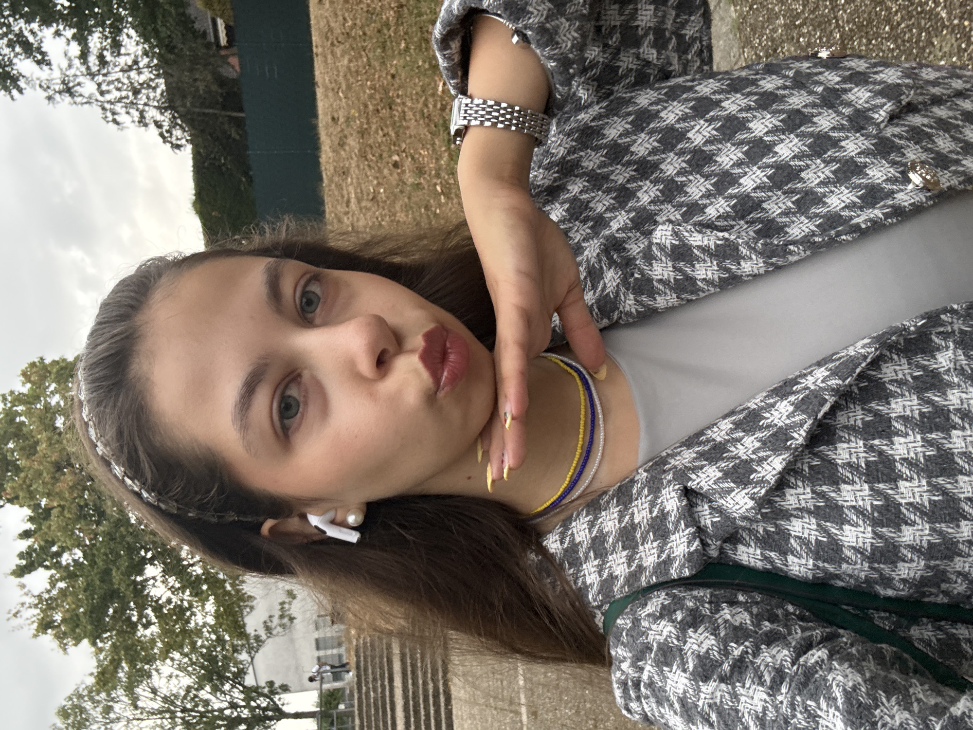 A young woman with long brown hair and a grey and white houndstooth jacket poses for a selfie outdoors. She is wearing a silver watch on her left wrist, white earbuds in her ears, and a beaded necklace. Her right hand is resting on her chin. The background shows trees, a grassy area, and a dark green fence. In the distance, there are stairs and two people walking. The lighting is overcast. The location is not specified in the image.