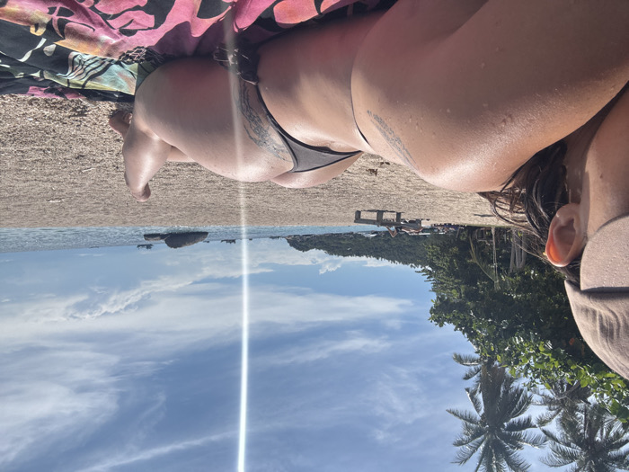 The image captures a daytime beach scene in Koh Tao, Thailand. In the foreground, an individual is observed lying prone on a multi-colored beach towel with a floral pattern, viewed from behind. The person wears a light-colored baseball cap and a dark-colored thong-style bikini bottom. Visible tattoos are present on the upper left shoulder and the left buttock/upper thigh. Moisture droplets are evident on the skin. The beach consists of fine sand mixed with small pebbles, extending towards the clear blue ocean. The left side of the scene is lined with lush green tropical vegetation, including multiple palm trees. In the midground, several individuals are visible further down the beach, some on lounge chairs. The background features several boats, including fishing vessels, anchored or moving in the ocean, alongside large rock formations in the water. Distant vegetated landmasses frame the horizon under a bright, partly cloudy sky. A vertical lens flare extends through the upper-middle portion of the image.
