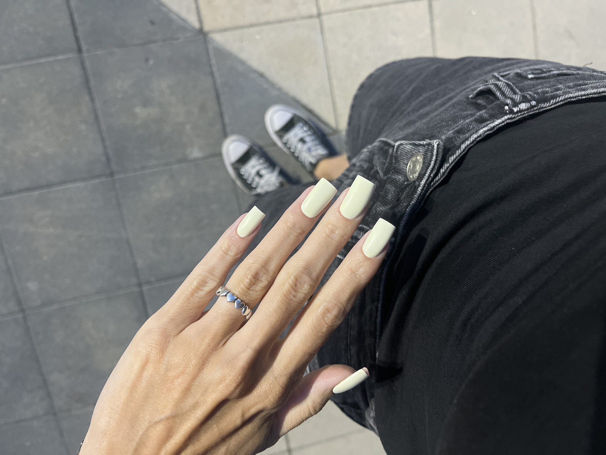 The image shows a close-up of a person's left hand and feet, wearing light yellow square-shaped artificial nails and a silver ring with a heart pattern. The person is wearing dark denim jeans and black and white Converse sneakers. The background appears to be an outdoor urban environment with gray tiled pavement. The image was taken in Nizhny Novgorod, Russia.