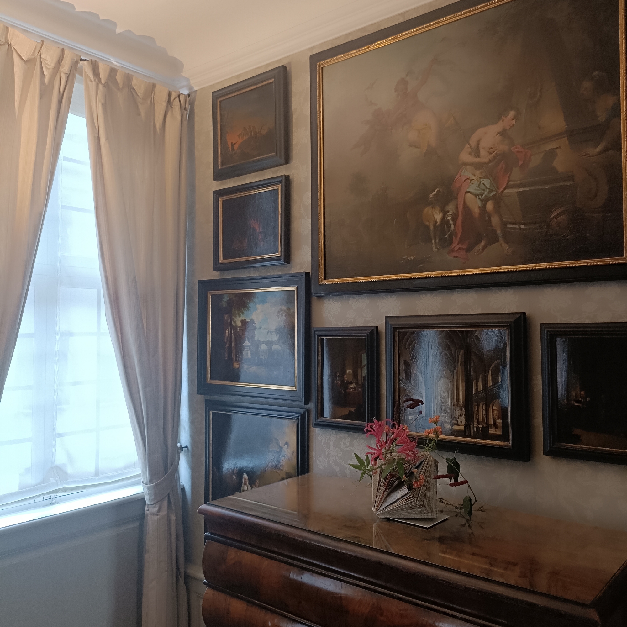The image depicts an interior scene, likely a room in a museum or a historic building, featuring several framed paintings on a wall. The paintings vary in size and subject matter, with one large central painting depicting a classical scene with figures and a dog. Smaller paintings are arranged around it, some showing landscapes and architectural scenes, and others darker and less discernible. The wall is covered in a patterned wallpaper. To the left of the paintings, sheer curtains are drawn over a window, allowing diffused light to enter the room. In the foreground, a polished wooden dresser or cabinet is visible, with a bouquet of flowers in a decorative holder placed on its surface. The lighting suggests an indoor setting with ambient light. There are no people clearly visible in the scene. This scene is located in the Städel Museum in Frankfurt, Germany.