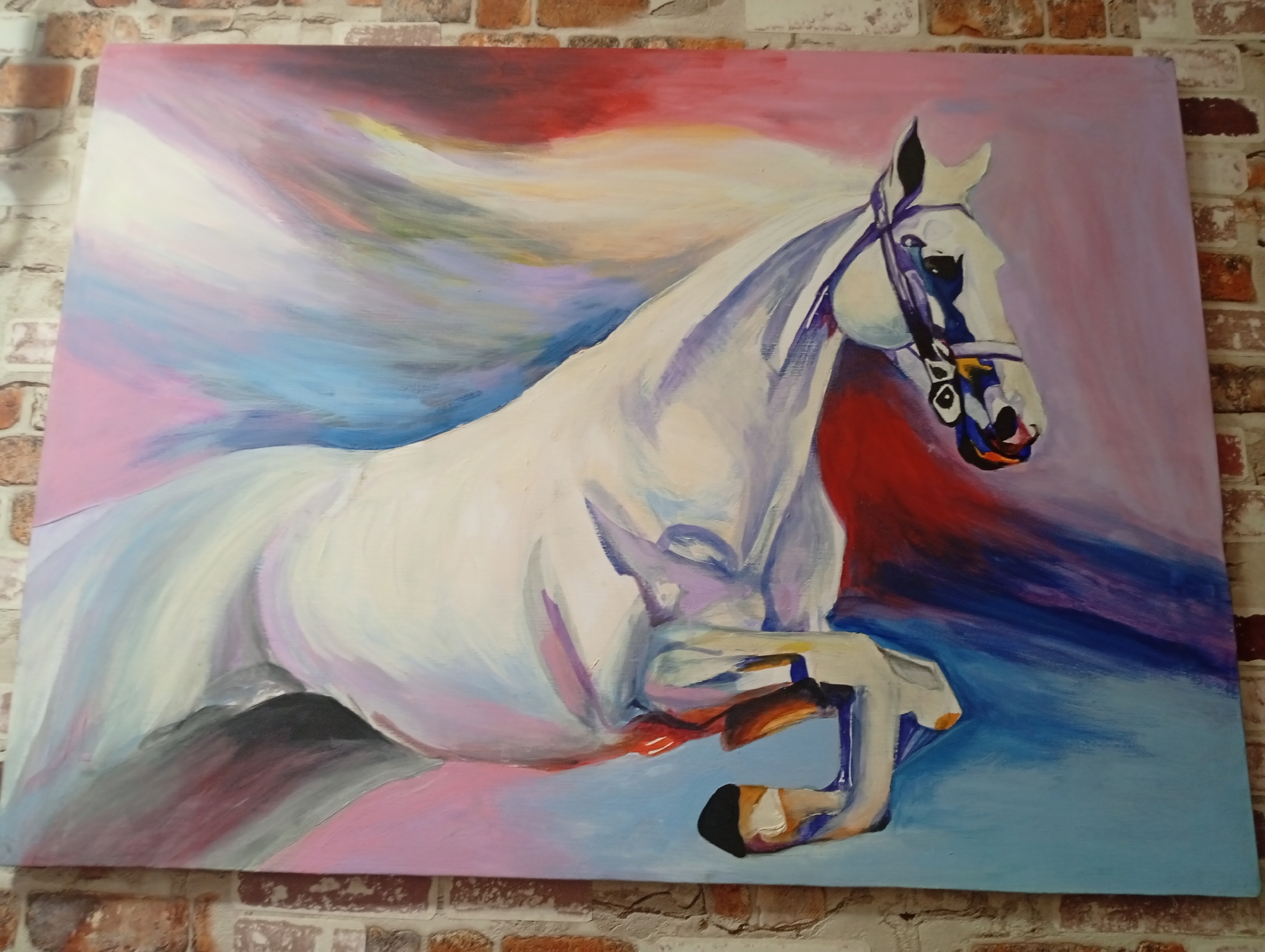 A vibrant, impressionistic oil painting depicts a white horse in motion against a colorful, abstract background. The horse, rendered with broad brushstrokes, dominates the canvas, its body a swirl of white, lavender, and hints of pink and blue. Its mane flows dramatically, a cascade of white and pastel hues. The background is a dynamic blend of reds, pinks, purples, blues, and yellows, suggesting movement and energy. The horse wears a bridle with blue and yellow accents. The painting is mounted on a brick wall, suggesting it is displayed in a home or gallery setting. The location is identified as Seýdi, Turkmenistan.