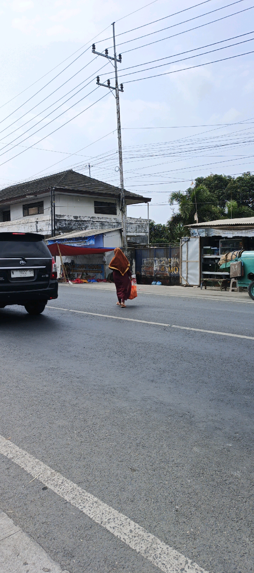 A medium shot captures a person walking away from the camera on a paved road in Kasiyan, Indonesia. The individual is wearing a dark maroon robe with a golden-brown headscarf and is carrying an orange plastic bag in their right hand. They are walking on the right side of the road, towards a row of low buildings and stalls.
In the foreground, the asphalt road features a white lane marking. A black SUV is visible on the left side of the frame, facing away from the camera. Behind the walker and to their left is a white building with a dark-tiled roof, featuring several windows. In front of this building is a stall with a blue and red tarp. To the right of the walker, there is a stall with a white facade and a metal corrugated roof, displaying shelves stocked with various items, including what appear to be glass tanks. A portion of a turquoise vehicle is visible to the far right.
The environment is characterized by utility poles and numerous overhead electrical wires crisscrossing the sky, which is overcast with light clouds. Trees are visible behind the stalls on the right. The scene depicts a typical street-level view with commercial activity and infrastructure common to a town or city.