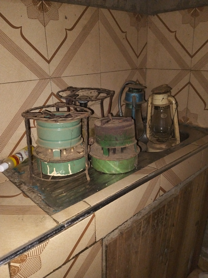 This image depicts an indoor utility area or kitchen within Nyansiongo ward, Kenya. The background wall is covered with light brown ceramic tiles featuring a repeating pattern of dark brown geometric lines and stylized floral motifs. A soiled metal or tiled countertop occupies the middle ground, with a visible metal sink basin located on the right.
Arranged on the countertop are several vintage items. From left to right, a prominent, heavily rusted dark metal framed kerosene or paraffin stove with a teal-green cylindrical fuel tank is visible. Next to it, a smaller, similarly rusted stove with a dark metal top and a green base is present. Partially obscured behind the leftmost stove is the rusted metal frame of another stove burner. Inside the sink basin, two vintage kerosene lanterns are placed. The left lantern features a blue top and a clear glass globe, while the right lantern is off-white with a clear glass globe displaying an "ANCHOR" brand label. On the far left of the countertop, a small yellow and white squeeze bottle with a white cap rests horizontally.
All metal objects show significant rust, dirt, and general wear. The countertop surface exhibits grime and reflective properties. Below the countertop, additional patterned tiles match the wall design, alongside a section of dark wooden paneling, possibly a cabinet door. No individuals are present in the scene.