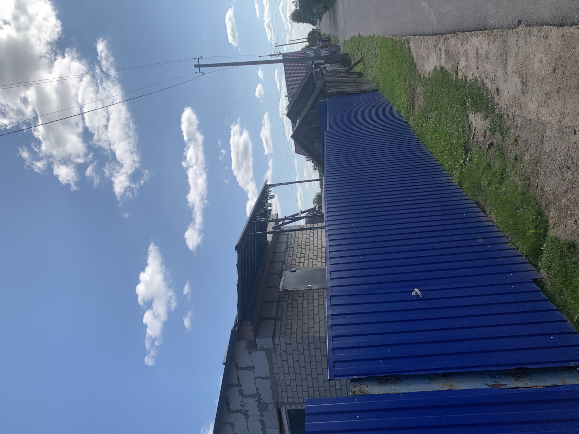 A vertical, eye-level, outdoor, daytime, full shot captures a rural street scene in Kapyĺ, Belarus. The foreground features a vibrant blue corrugated metal fence running diagonally from the lower left to the middle right of the frame. To the right of the fence is a narrow strip of green grass, followed by a dirt path, and then a gray asphalt road that recedes into the distance.
Beyond the fence, construction is evident on a single-story building, partially constructed with light-colored bricks and cement blocks. A dark metal awning extends from the building, supported by posts. A silver door is visible on the side of the building. Further down the street, a few more buildings with red roofs and older wooden structures are visible.
Overhead, a bright blue sky is dotted with scattered white clouds. Power lines stretch across the sky, supported by wooden utility poles. The overall impression is one of a quiet, rural community on a clear day.