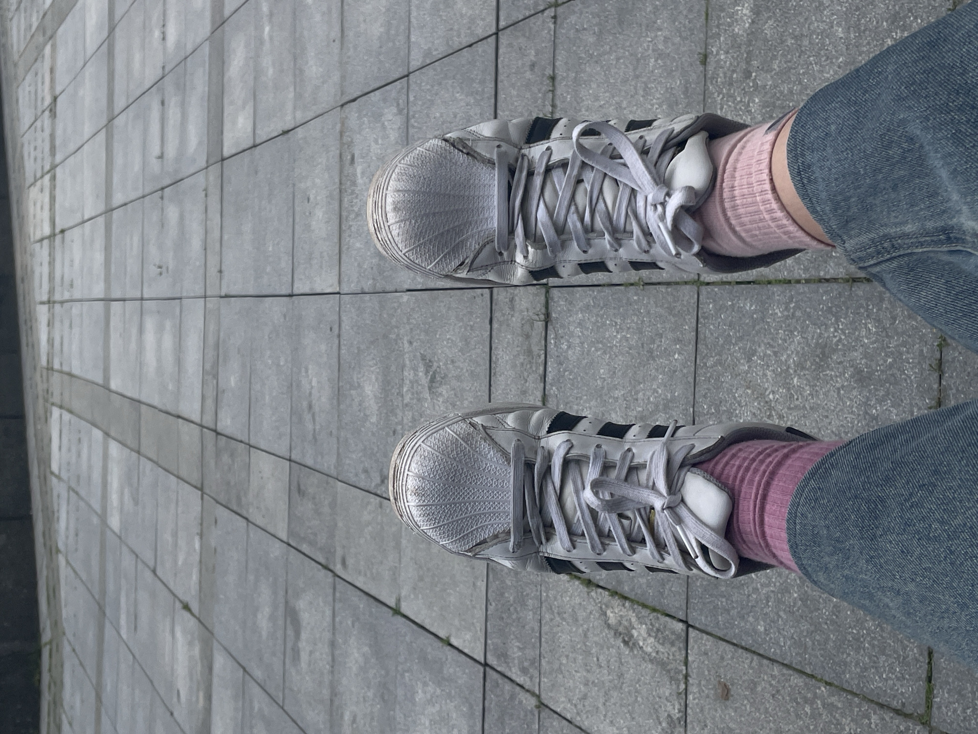 This media file shows a top-down view of a person's feet wearing white Adidas sneakers with black stripes, pink socks, and blue denim jeans, standing on a sidewalk paved with gray rectangular tiles. The shoes appear well-worn, with some scuff marks and creasing on the toe boxes. The socks are ribbed and extend above the ankle, peeking out from the top of the sneakers. The jeans are a classic blue denim. The setting is an outdoor urban environment, likely a city sidewalk, indicated by the tile paving and the suggestion of a building edge or wall to the left of the frame.
The lighting suggests it is daytime, with no strong shadows indicating a specific time of day, but the overall light is soft, possibly overcast or diffused. There is no visible text in the image. The image focuses on the footwear and lower legs of the individual, implying an activity of walking or standing in an urban area. The composition is simple, with the feet centered in the frame and the patterned sidewalk providing a textured background. There are no clear indicators of emotion or a specific event taking place, presenting a common, everyday scene. The location context provided is Moscow, Russia, which aligns with the urban setting.