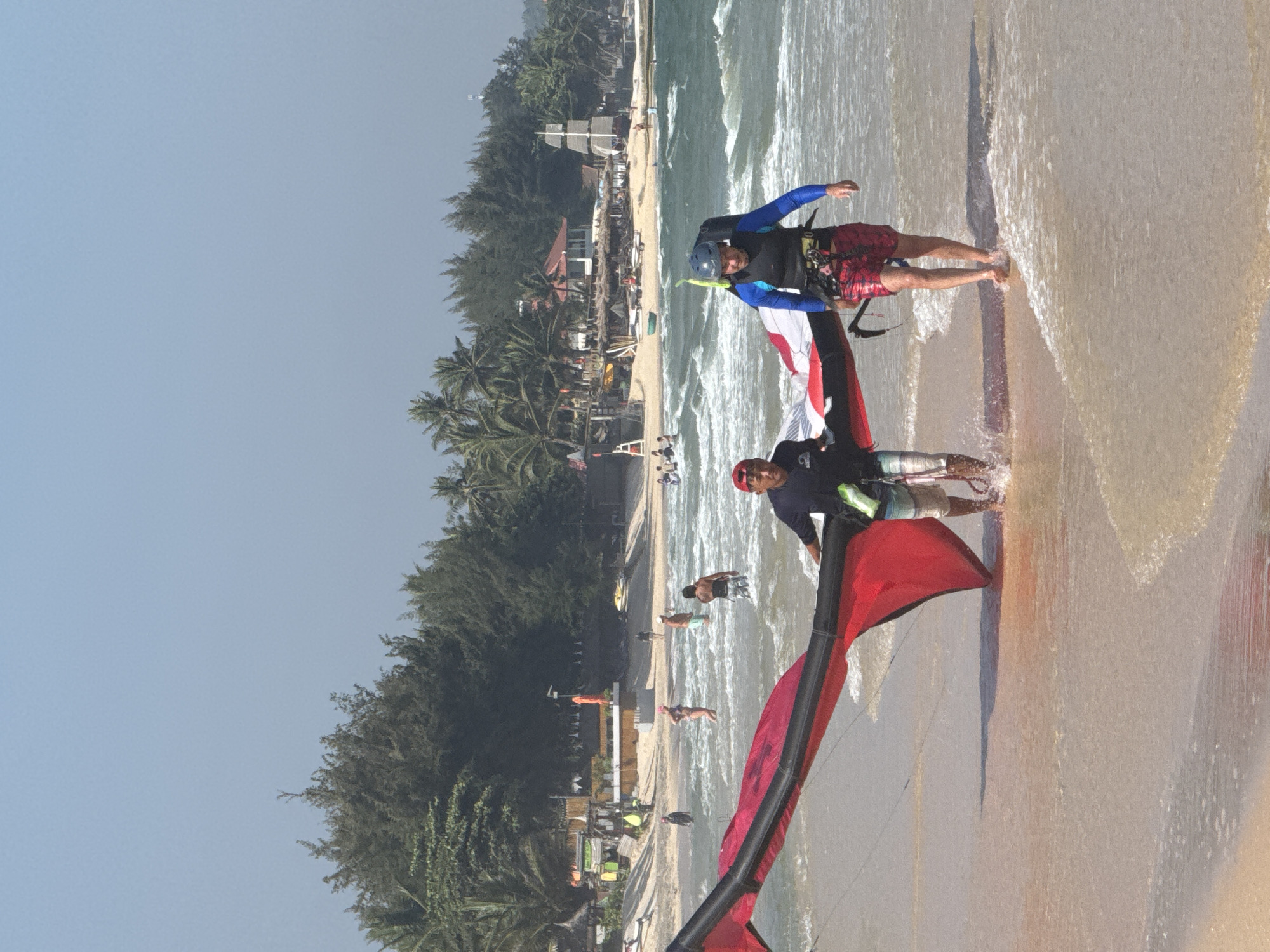Two men are walking in the shallow surf at a beach in Mũi Né, Vietnam. They are carrying a large red and black kite, likely for kitesurfing. The man on the left wears a dark blue shirt, shorts, and a red cap. The man on the right wears a blue long-sleeved top, a life vest, red and black patterned shorts, and a helmet. In the background, there are trees, buildings, and other people on the beach and in the water. The water is calm with small waves.