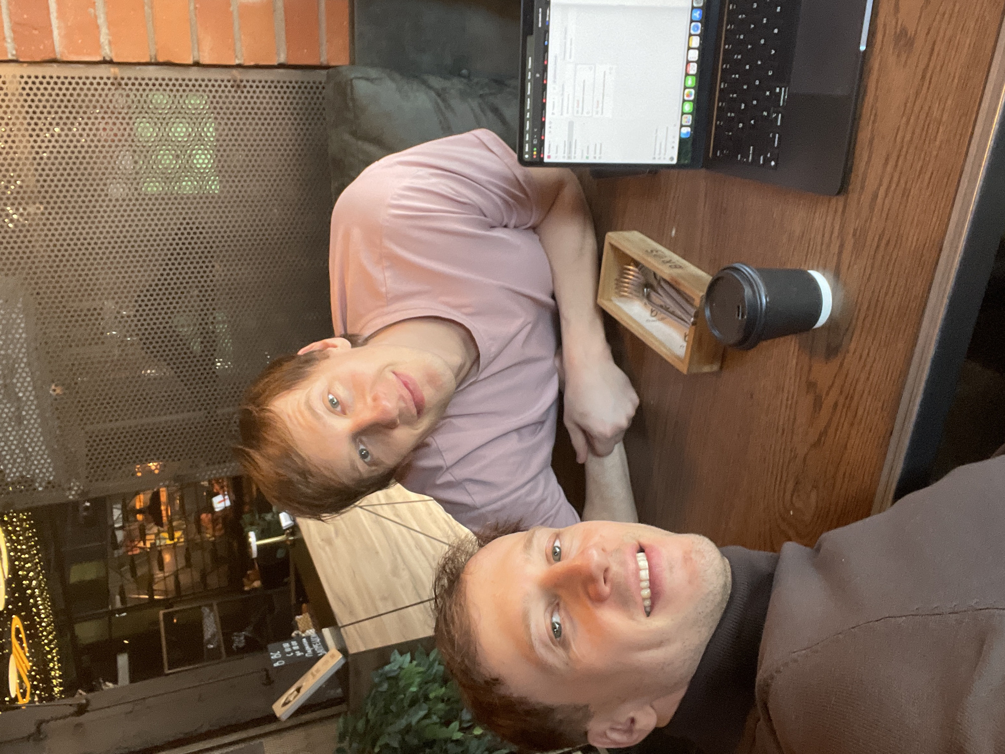 Two Caucasian males are positioned in close proximity, likely at a table in a public space. One man is wearing a light pink t-shirt and the other a brown turtleneck sweater. A laptop sits open on the table. Nearby are a cup with a lid and a wooden box containing eating utensils. The location appears to be Saint Petersburg, Russia, based on external environmental details visible through windows in the background.