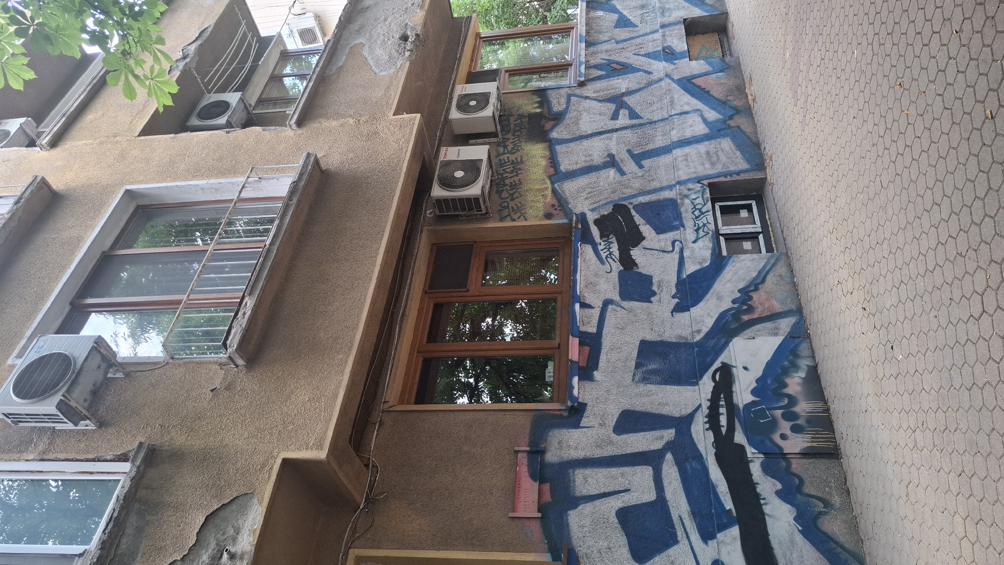 The image depicts the exterior of a multi-story building in Sofia, Bulgaria, characterized by its weathered facade and extensive graffiti. The building's exterior walls are painted a light brown, with noticeable patches of peeling plaster and exposed concrete. Several windows are visible on different levels, some with wooden frames and others with white frames. Air conditioning units are mounted on the exterior walls, with two units prominently placed side-by-side on one level.
The lower section of the building, at street level, is heavily adorned with blue and white graffiti, featuring large, stylized lettering and abstract shapes. A portion of the graffiti appears to cover a metal utility panel or door. There are also two small, recessed windows with white frames at the base of the building, partially obscured by the graffiti.
The ground in front of the building is paved with hexagonal interlocking concrete tiles. A few fallen leaves are scattered on the pavement. Green foliage from trees overhangs the upper portion of the building, suggesting a tree-lined street or courtyard. The overall impression is one of urban decay mixed with vibrant street art. There are no people visible in the image.