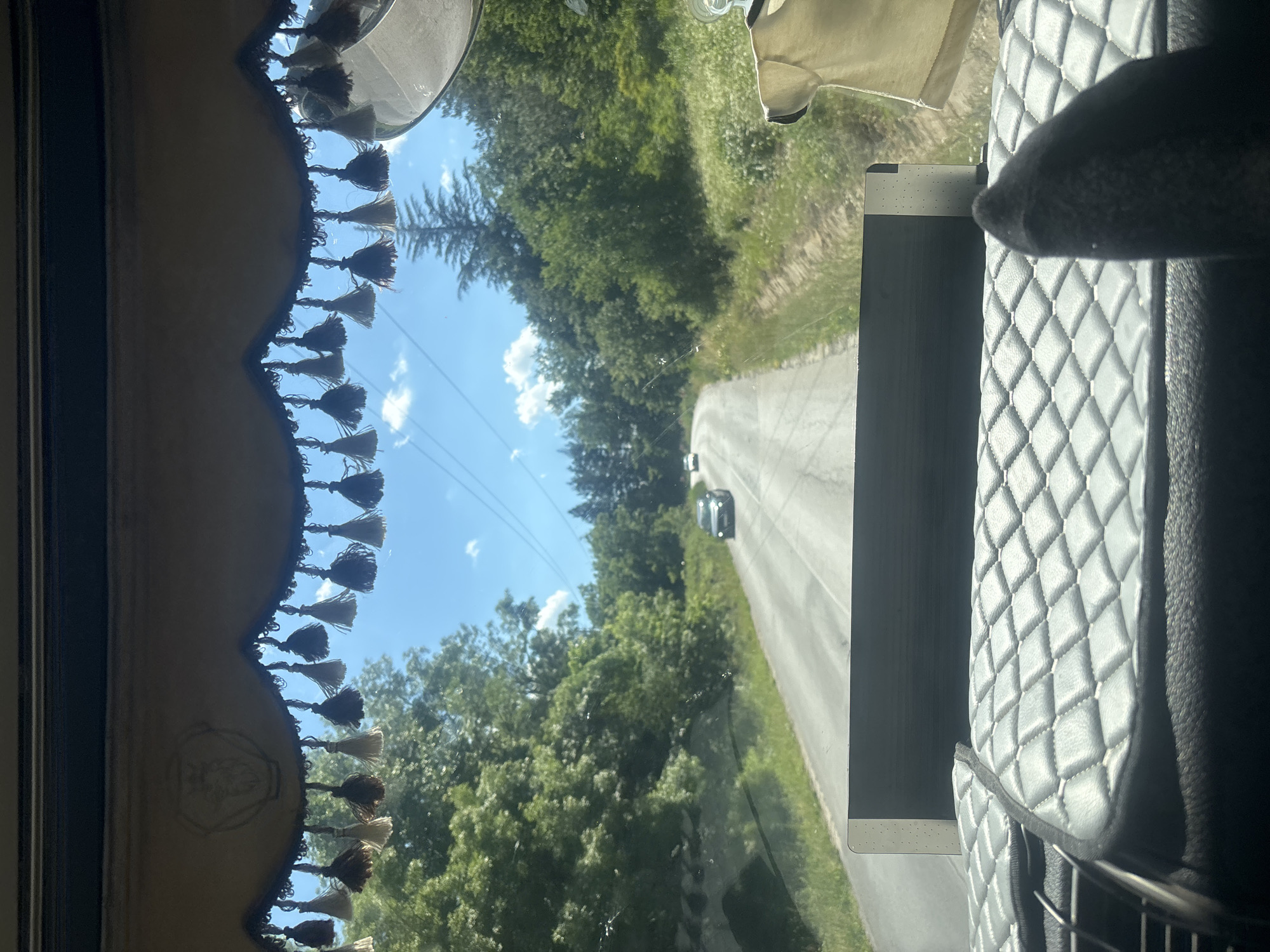 The image, taken from inside a vehicle in Topolovgrad, Bulgaria, captures a view through the windshield. A decorative trim with numerous tassels hangs across the top of the windshield. The road ahead is paved and curves to the left, bordered by lush green trees and vegetation. Two cars are visible on the road in the distance. The sky is blue with scattered white clouds, and power lines run overhead. In the foreground, the interior of the vehicle is visible, with a quilted dashboard and what appears to be a black sock or fabric in the lower right corner. A beige garment is also hanging from the right side of the frame.