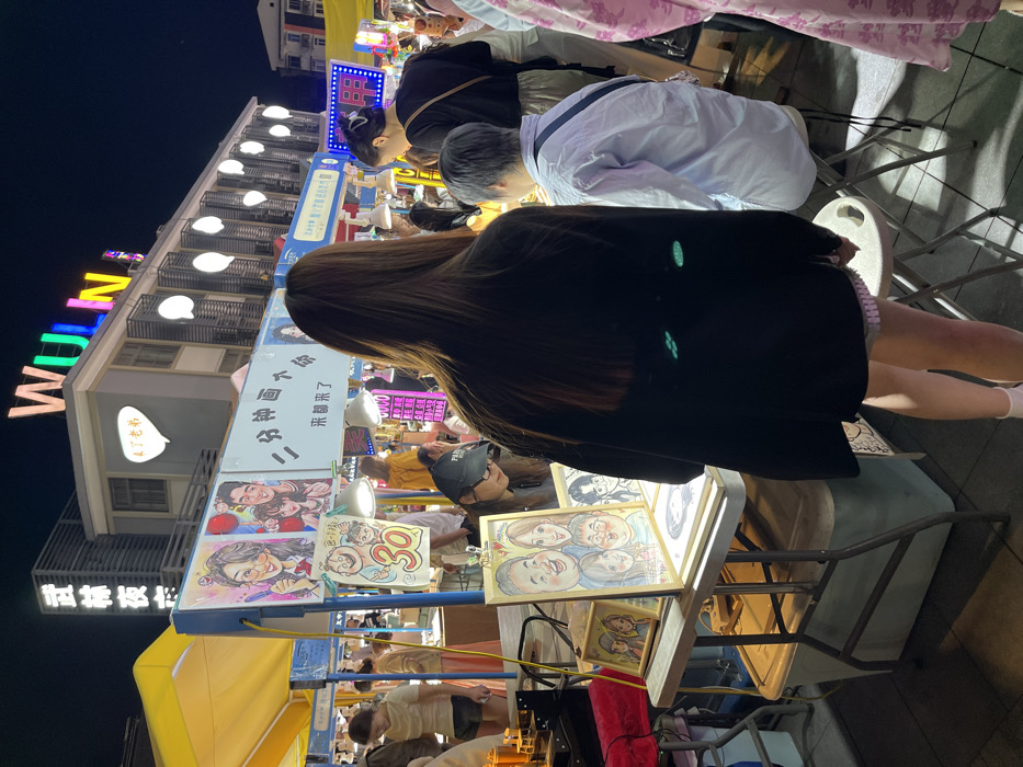 The image shows a street scene in Hang Zhou Shi, China, at coordinates 30.264 latitude and 120.156 longitude. It's a night market with various vendors, including a caricature artist who is drawing a portrait of a man. The artist is sitting at a table with other drawings displayed. A woman with long dark hair is standing by the table, seemingly observing the artist's work. Behind her is a man wearing a light blue shirt, also observing the process. The scene is dimly lit, and the atmosphere is casual and relaxed. The building in the background has a sign that reads "WULN". There are many other people walking around in the night market, but they are not the focus of the image. The focus is on the artist, the woman, and the man.