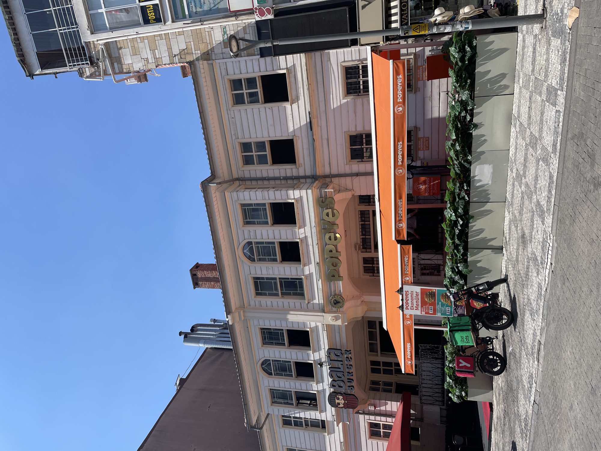 The image captures a street scene in Kadıköy, Turkey, on a clear, sunny day. In the foreground, a cobblestone street is visible, with a parked delivery motorcycle on the right side. The motorcycle is equipped with two large green delivery boxes, one labeled "Uber Eats" in white letters, and a smaller pink box attached to the back. The motorcycle has a rider dressed in dark clothing.
Dominating the mid-ground is a white, two-story building housing two businesses: "Bağla Burger" on the left and "Popeyes" on the right. "Bağla Burger" features a distinctive logo with a stylized illustration of a man's face and the name "Bağla Burger" in blue lettering. "Popeyes" is marked by its prominent orange awning and the restaurant's name displayed in large, golden letters. Under the awning, there is a menu board advertising Popeyes "Ekonomix Menüler" (Economix Menus). A line of potted green plants separates the businesses from the sidewalk.
The building itself has a traditional architectural style with white wooden siding and several arched windows on the upper floor, some of which are open. Metal pipes and a brick chimney are visible on the roof.
To the right of the Popeyes establishment, a multi-story building with a stone facade and modern windows is visible, featuring a "POSTIJ" sign. A lamppost stands on the sidewalk next to this building. The sky is a clear, vibrant blue, indicating fair weather.