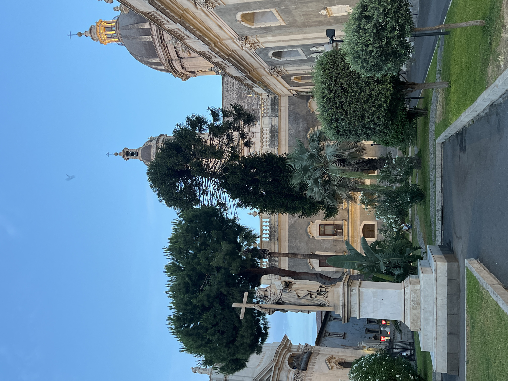 The image depicts a twilight scene in Piazza Duomo, Catania, Italy, featuring the Catania Cathedral. In the foreground, a white marble statue of a woman holding a cross and a book stands on a tiered pedestal. Behind the statue, a large pine tree and a palm tree are visible, with manicured bushes and green grass in the immediate vicinity. The Catania Cathedral's imposing facade and dome dominate the background, with architectural details such as arched windows and ornate carvings visible. The sky is a clear blue, transitioning to dusk. A faint bird is captured in flight against the sky. The lighting suggests a late afternoon or early evening setting, with the warm glow from the cathedral dome indicating artificial illumination beginning to activate. A person is seated in the background, in the grassy area. The overall atmosphere is serene and historic, characteristic of a European piazza.