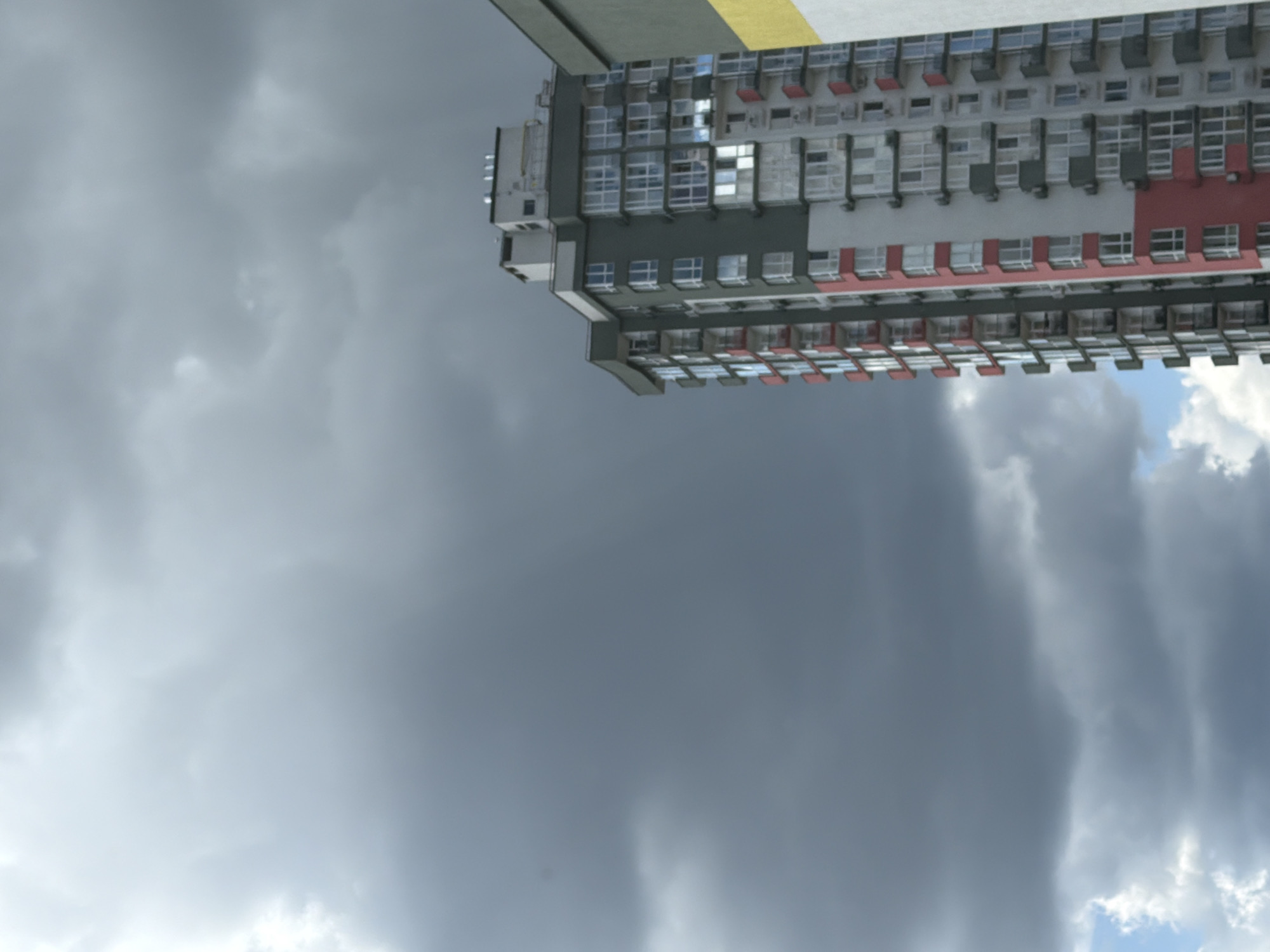 The image shows a tall apartment building with a modern facade, featuring a mix of white, grey, and red sections, and numerous balconies with glass railings. The building is partially obscured by a dark, cloudy sky, suggesting overcast weather. To the right of the main building, another structure with a yellow and green exterior is visible. The overall scene depicts an urban environment, likely a residential area in Kyiv, Ukraine, under a stormy sky. There are no people visible in the image.