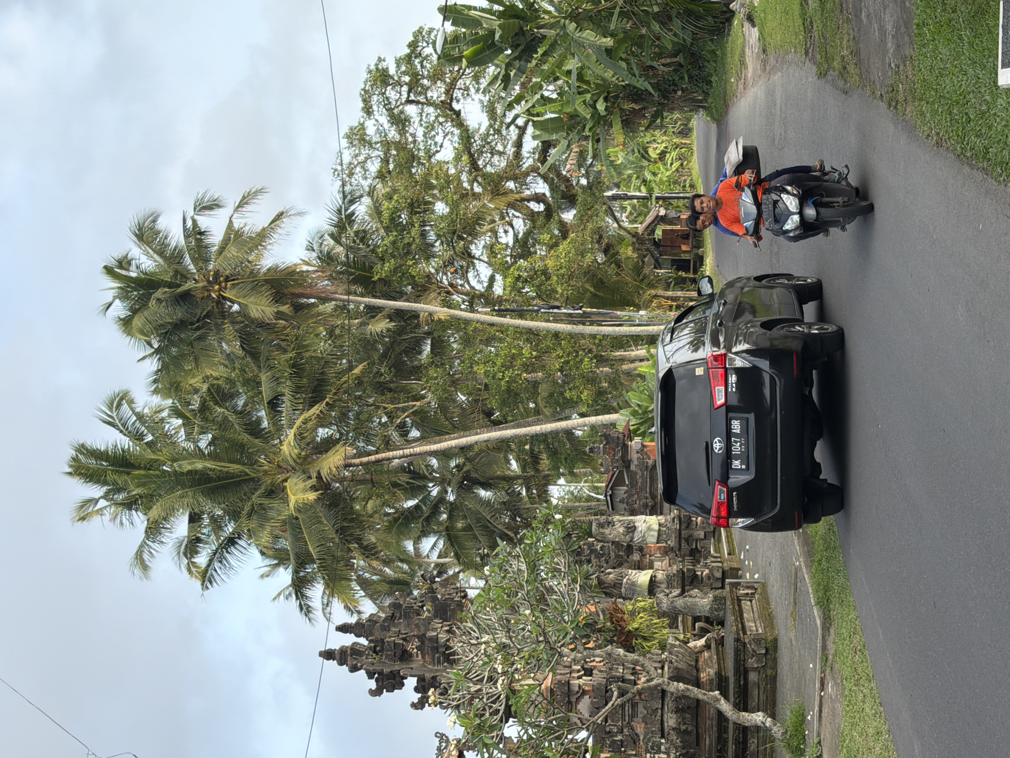 The image depicts a roadside scene with lush tropical vegetation and traditional Balinese architecture. A black SUV, identified by its license plate "DK 1047 ABR", is parked on the right side of the asphalt road. In front of the SUV, two men are riding a black motorcycle. The driver, wearing an orange shirt, is looking forward, while the passenger, in a blue shirt, is looking towards the camera. They are carrying a large, flat object wrapped in paper on the back of the motorcycle.
To the left of the road, a large, intricately carved stone structure, likely a temple gate, stands amidst dense greenery. Several tall palm trees dominate the background, with their fronds reaching towards a cloudy sky. The road curves into the distance, flanked by more trees and foliage, suggesting a rural or suburban setting. The lighting suggests it is daytime, possibly late afternoon, given the soft light.