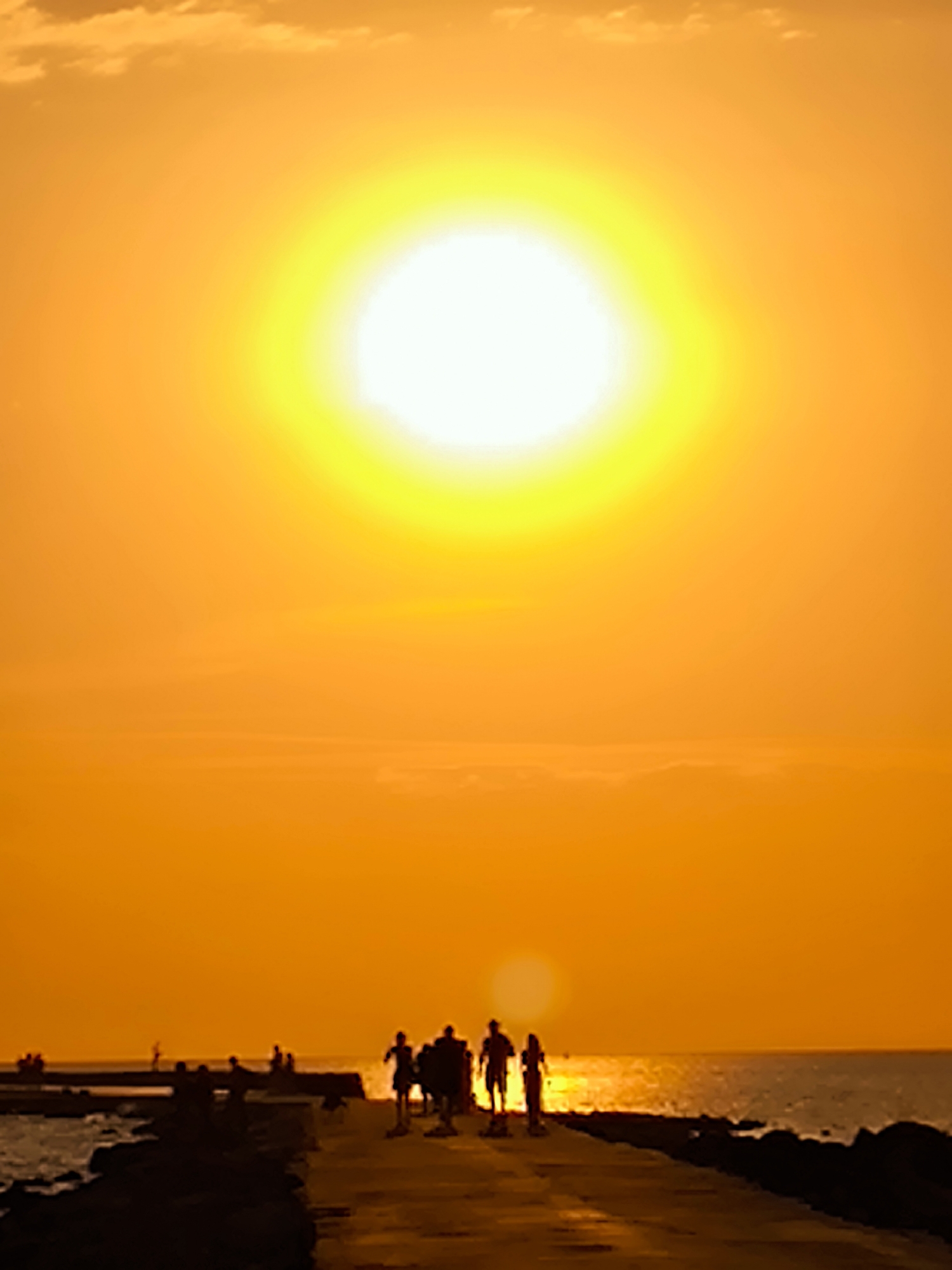 This is a vertical, slightly tilted, color photograph of a sunset over a body of water, likely the sea or a large lake, with a promenade in the foreground. The sun is a large, bright, yellow-white orb dominating the upper center of the frame, casting a warm orange glow across the entire scene. Wispy clouds are visible in the upper left corner, tinged with orange from the setting sun.
In the mid-ground, a paved promenade stretches horizontally across the image, silhouetted against the bright sky. Several figures, appearing as dark silhouettes, are walking along the promenade. There are at least five individuals clearly visible in a group on the right side of the promenade, with others potentially further down. Their postures suggest movement, with some appearing to be walking or running. The figures are small relative to the vastness of the sky and sun, emphasizing the scale of the natural light display.
To the left of the promenade, dark rocks are visible along the water's edge. The water itself reflects the orange and yellow hues of the sky, creating a shimmering surface. The overall atmosphere is tranquil and serene, characteristic of a sunset by the water. There is no explicit information within the image to indicate a specific location in Riga, Latvia.