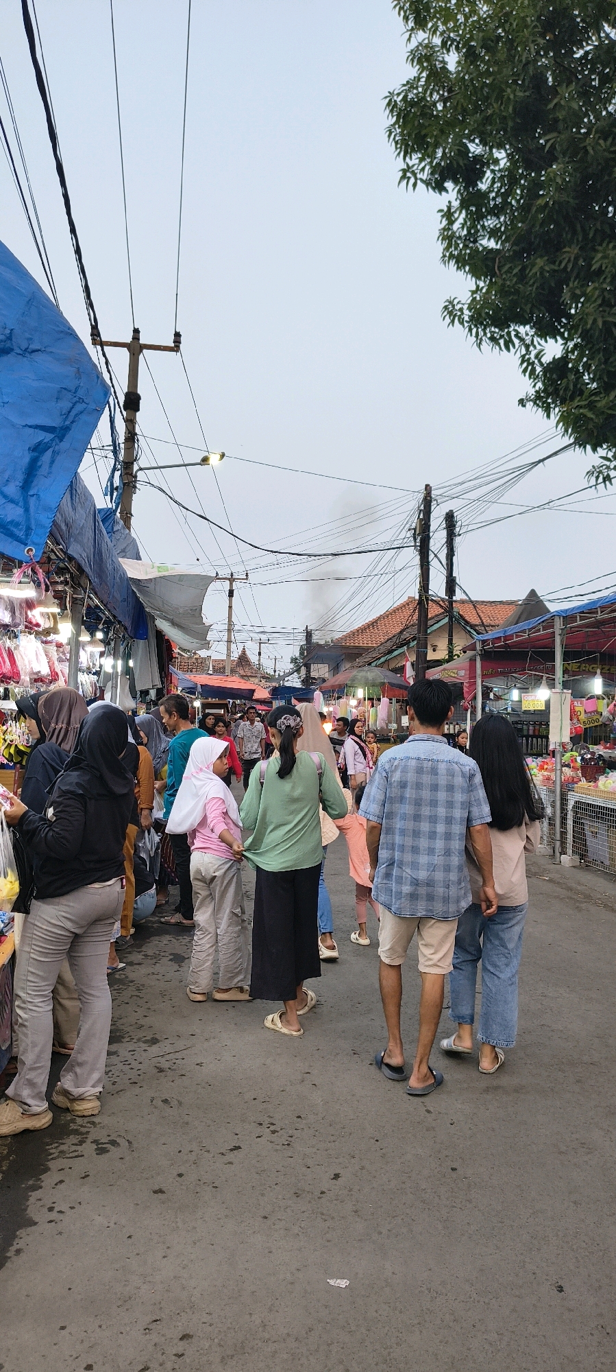 This image depicts a bustling outdoor market scene in Trusmi Wetan, Indonesia, likely during the late afternoon or early evening, given the overcast sky and artificial lighting from the stalls. A crowd of people, including men, women, and children, are walking along a paved pathway lined with market stalls. The stalls are covered with blue and white tarpaulins, displaying various goods such as clothing and accessories. Several individuals are seen browsing the merchandise, with some holding shopping bags. Prominent in the foreground are a man in a blue plaid shirt and light-colored shorts, and a woman in light blue jeans, walking away from the camera. To their left, a young girl in a pink shirt and white hijab stands next to a woman in a green top and black pants. In the background, more market stalls and a few buildings with terracotta roofs are visible, along with a dense network of overhead electrical wires and utility poles. A light mist or smoke is visible in the distance, suggesting potential cooking or industrial activity. The ground is a dark, damp pavement with some scattered debris. The overall atmosphere is one of everyday commerce and community gathering in a semi-urban setting.