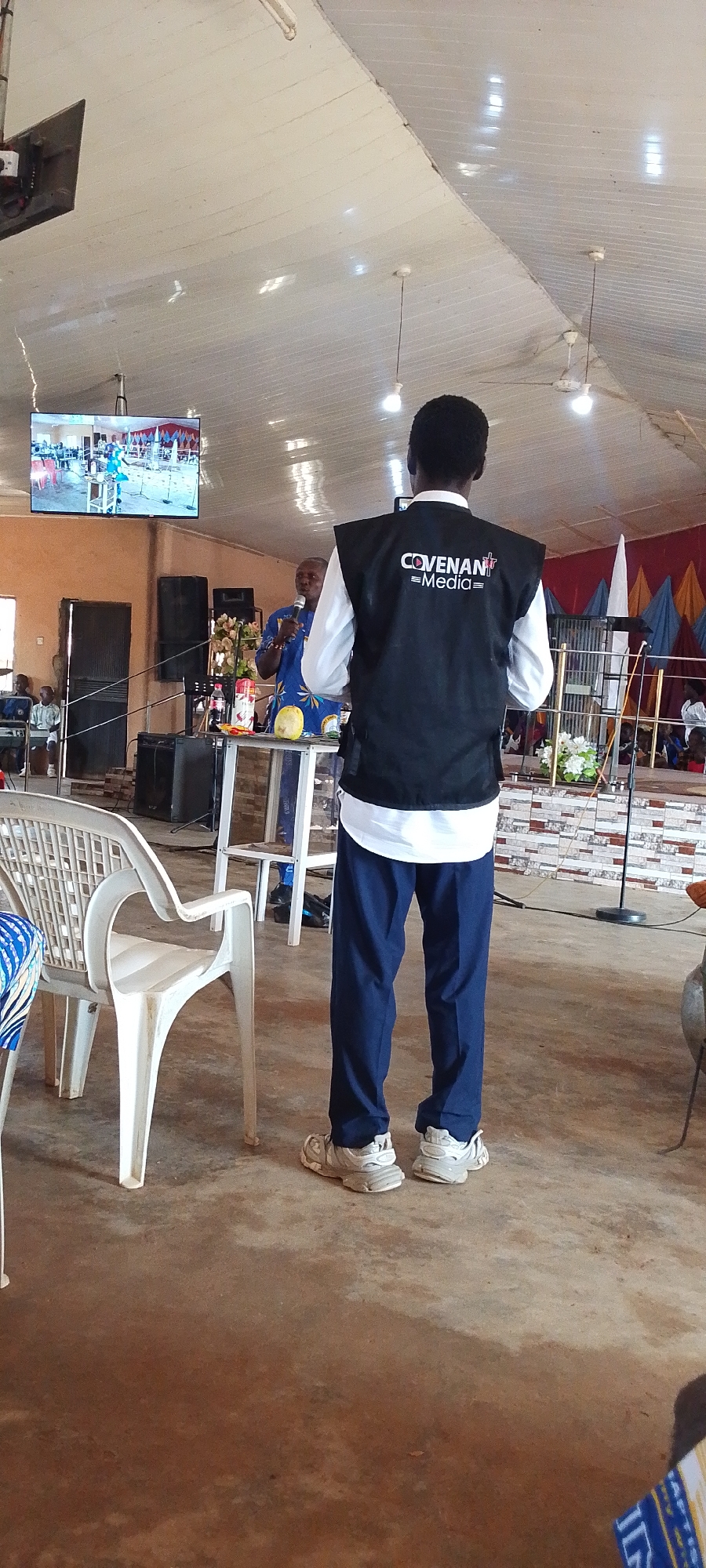 This media file depicts an indoor scene, likely a church or a community hall, in Gumel, Nigeria. The setting is characterized by a light-colored, sloped ceiling with exposed lighting fixtures and ceiling fans. In the foreground, a person from behind is wearing a black vest with "COVENANT Media" text visible on the back, over a white long-sleeved shirt and blue trousers. They are also wearing white athletic shoes.
To the left, a white plastic chair is partially visible. In the mid-ground, a man in a blue patterned shirt is speaking into a microphone, standing behind a table with drinks and a yellow fruit. Behind him, there are large black speakers and decorative flowers. A large television screen is mounted on the wall above, displaying an image of people on a stage, possibly a performance or service. The overall atmosphere appears to be that of a gathering or event, likely during the daytime given the interior lighting. The floor is concrete. Further back, seating and a stage area with colorful drapes are visible.