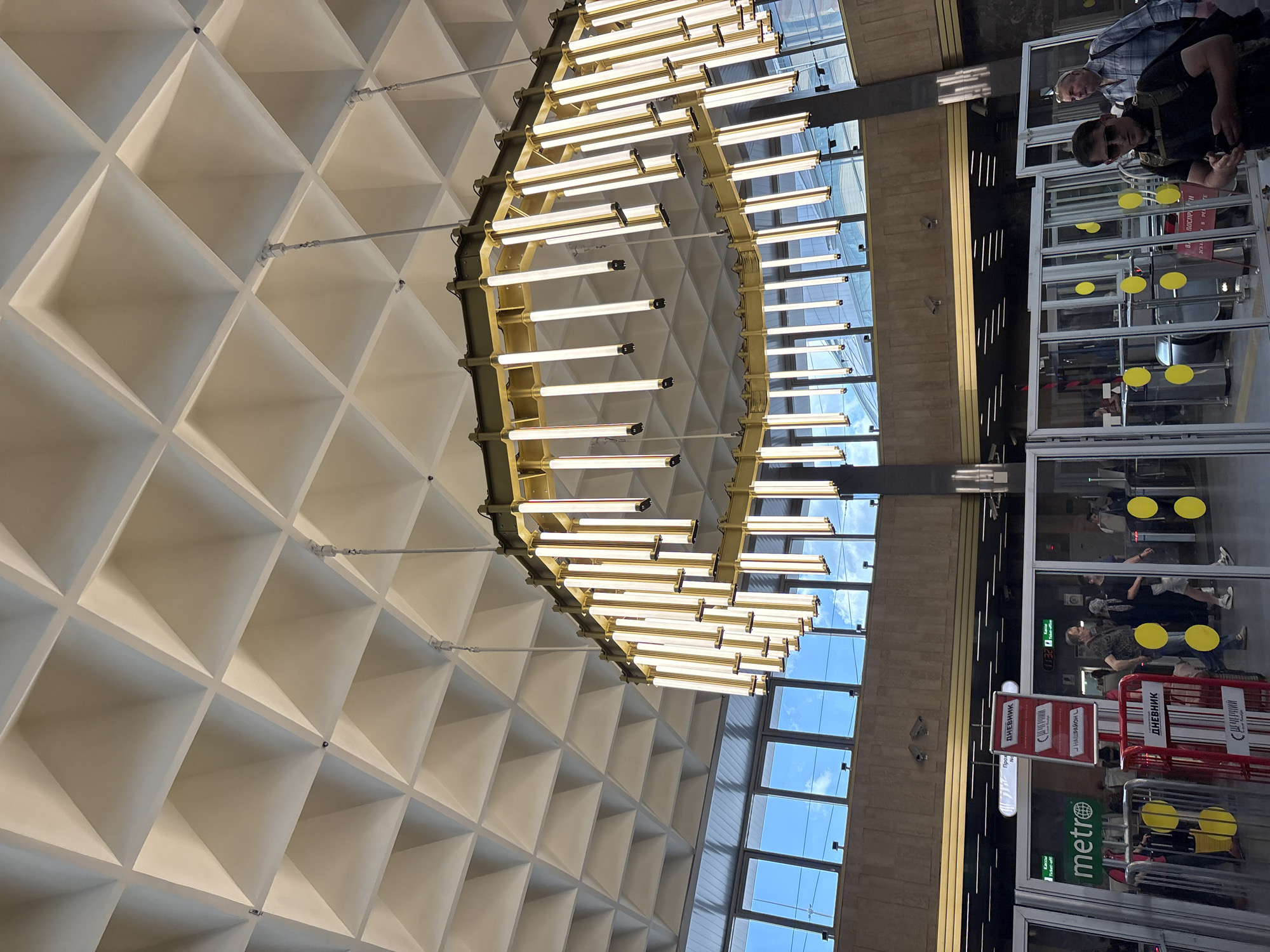 The image shows the interior of a metro station in Saint Petersburg, Russia. The station features a distinctive coffered ceiling and a large, modern chandelier with multiple fluorescent lights. Large windows at the top allow natural light to filter in, revealing a blue sky with some clouds. Below the windows, a beige tiled wall with a golden trim runs horizontally. In the foreground, there are several glass turnstiles for entry and exit. A red metal barrier with yellow circular stickers is also visible. Several people are seen passing through the turnstiles or standing nearby. One person in the foreground is wearing sunglasses and a backpack, holding a smartphone. A man in a plaid shirt is walking in the background. Signs indicating "metro" and Russian text are visible on the turnstiles and barriers. The overall impression is a well-lit, contemporary public transportation hub.