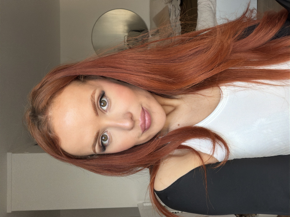 A headshot-style image captures a woman indoors, located in a residence in Paris, France. The subject, a woman with long, auburn hair and brown eyes, gazes directly at the camera. She has a light complexion, defined eyebrows, and wears visible eye makeup and light pink lipstick. Her attire consists of a white ribbed tank top, with a black garment covering her left shoulder and arm. The background features light-colored walls. A round wall-mounted mirror is visible on the right side, reflecting ambient light. Below the mirror, a partially visible bed or sofa with light-toned bedding is present.