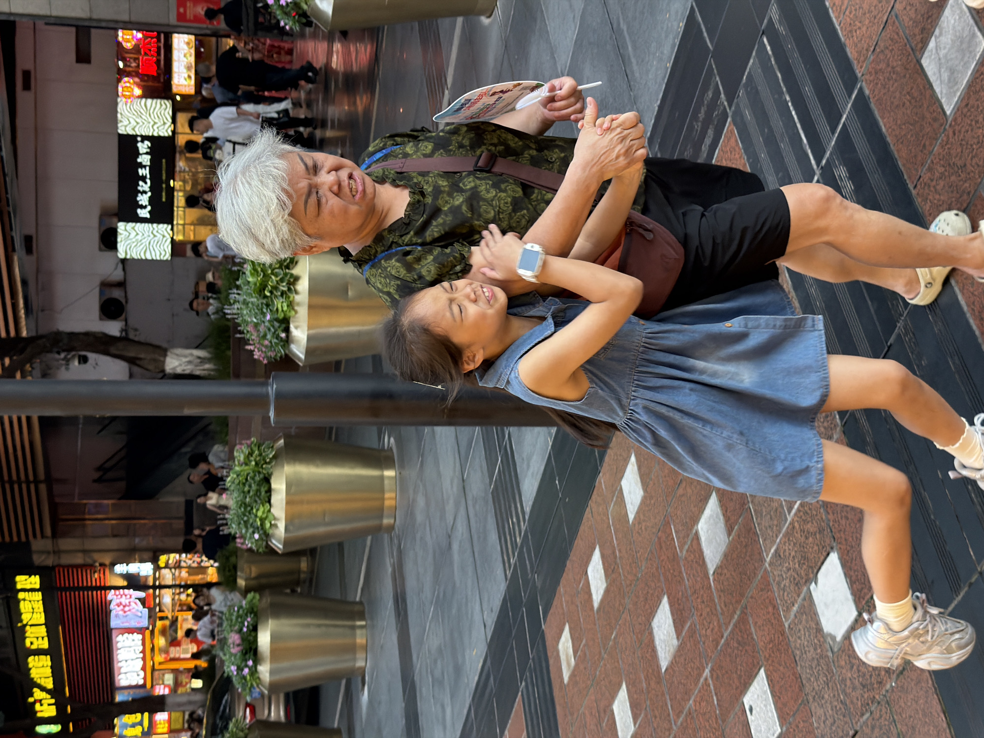 A young girl in a denim dress is being pulled by an older woman with white hair, who is wearing a green floral shirt and black shorts. The girl is smiling and laughing, while the woman appears to be speaking or yelling, her mouth open and eyes closed. The woman is holding a white fan with a colorful design. The girl is wearing white socks and silver sneakers, and the woman is wearing white clogs. They are standing on a tiled sidewalk, with planters and buildings in the background. The scene is outdoors, possibly in a shopping district, with neon signs visible in the distance. The time of day appears to be late afternoon or early evening, given the lighting. The atmosphere seems lively and playful. The text on the neon signs is in Chinese.