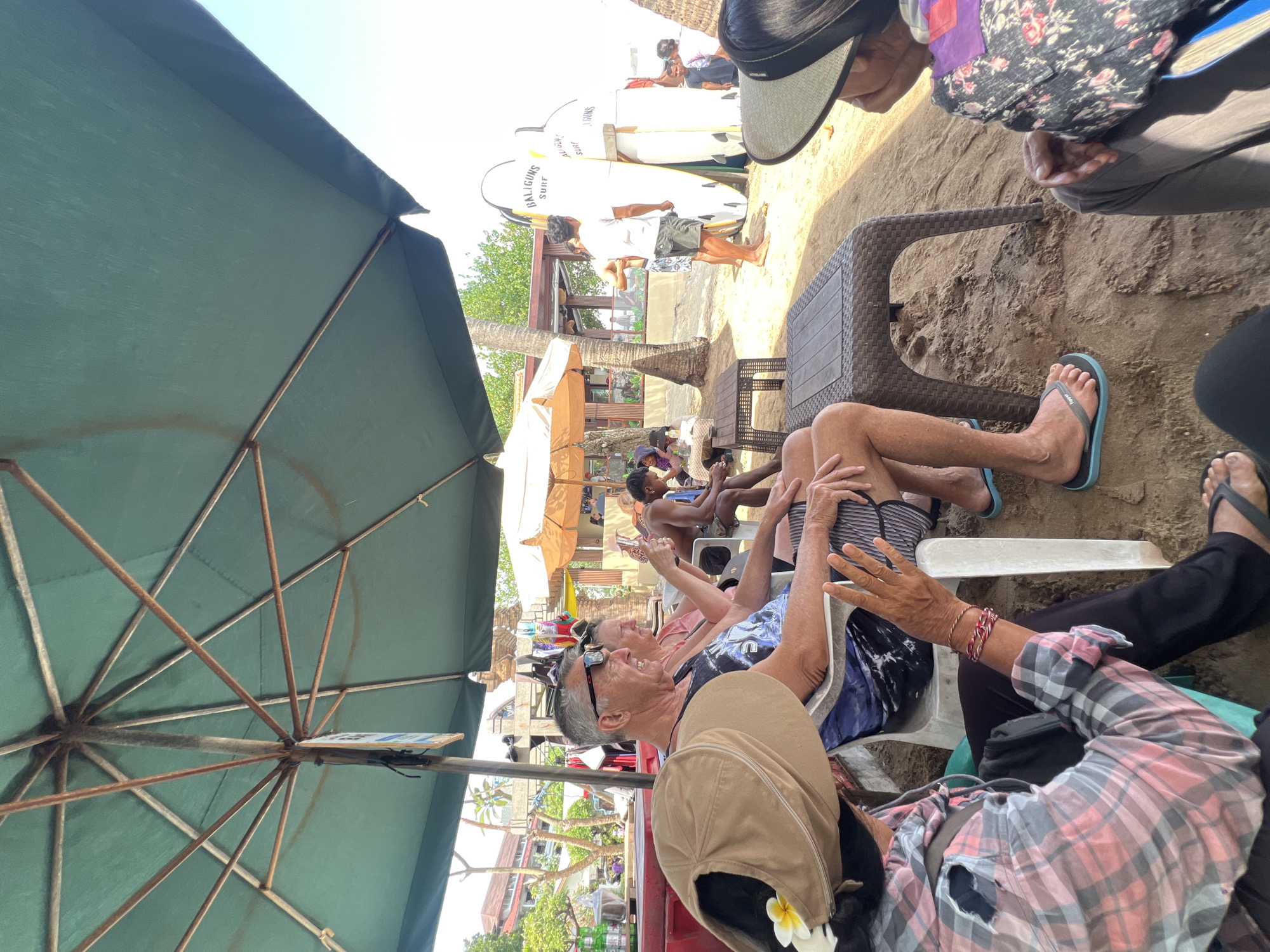 In Kuta, Indonesia, on a beach, a group of people are seated under a large green umbrella. In the foreground, a person wearing a tan hat with a yellow flower is seated, with their arm extended. To their right, a man with sunglasses and a patterned tank top is sitting, looking to his right and smiling. Next to him, a woman in a pink shirt is also seated. Further to the right, a shirtless young man with dark skin is seated, looking down. In the background, there are surfboards with "BALIGUNS SURF" written on them, a beach restaurant, and palm trees. The ground is covered in sand.