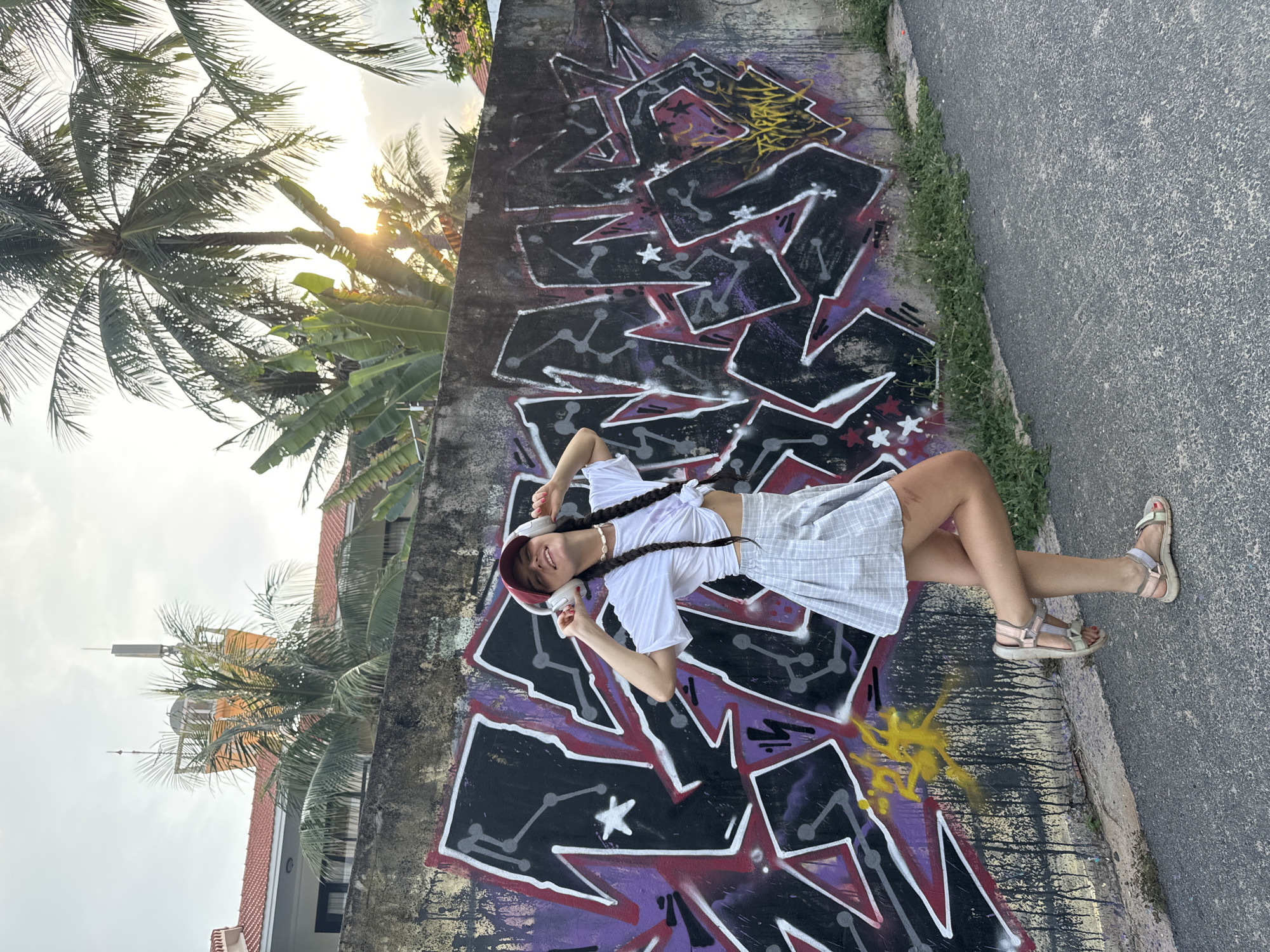 A young woman stands in front of a graffiti-covered wall in Phu Thuy Ward, Vietnam. She is wearing a white crop top, a grey plaid skirt, a red baseball cap, and white headphones, with her hands touching the headphones. She has a happy expression, with her eyes closed and her head tilted back. Her hair is styled in two braids. She is wearing sandals and a shell necklace. The wall behind her is covered in black and purple graffiti, with some yellow spray paint. Palm trees and banana trees are visible behind the wall, along with a building and cell towers in the background. The sun is setting, casting a warm glow on the scene.
