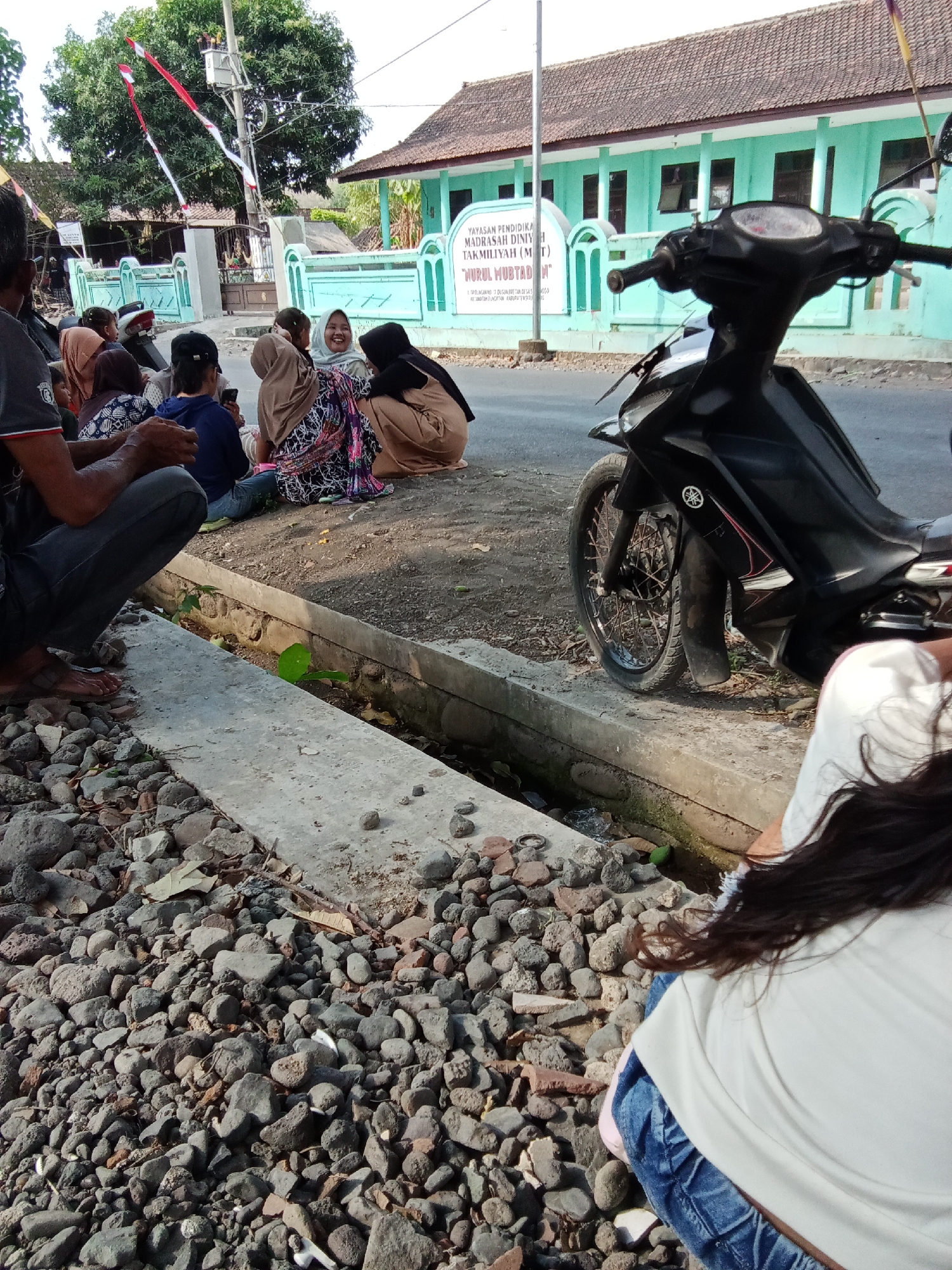 In Trebungan, Indonesia, a group of individuals, including men, women, and children, are gathered outdoors on a dirt and gravel surface next to a concrete drainage ditch. They are seated or squatting, appearing to be in a casual social setting. A black motorcycle is parked nearby. In the background, a light turquoise building with a brown tiled roof is visible, with a sign bearing Indonesian text. The environment suggests a rural or semi-urban area.